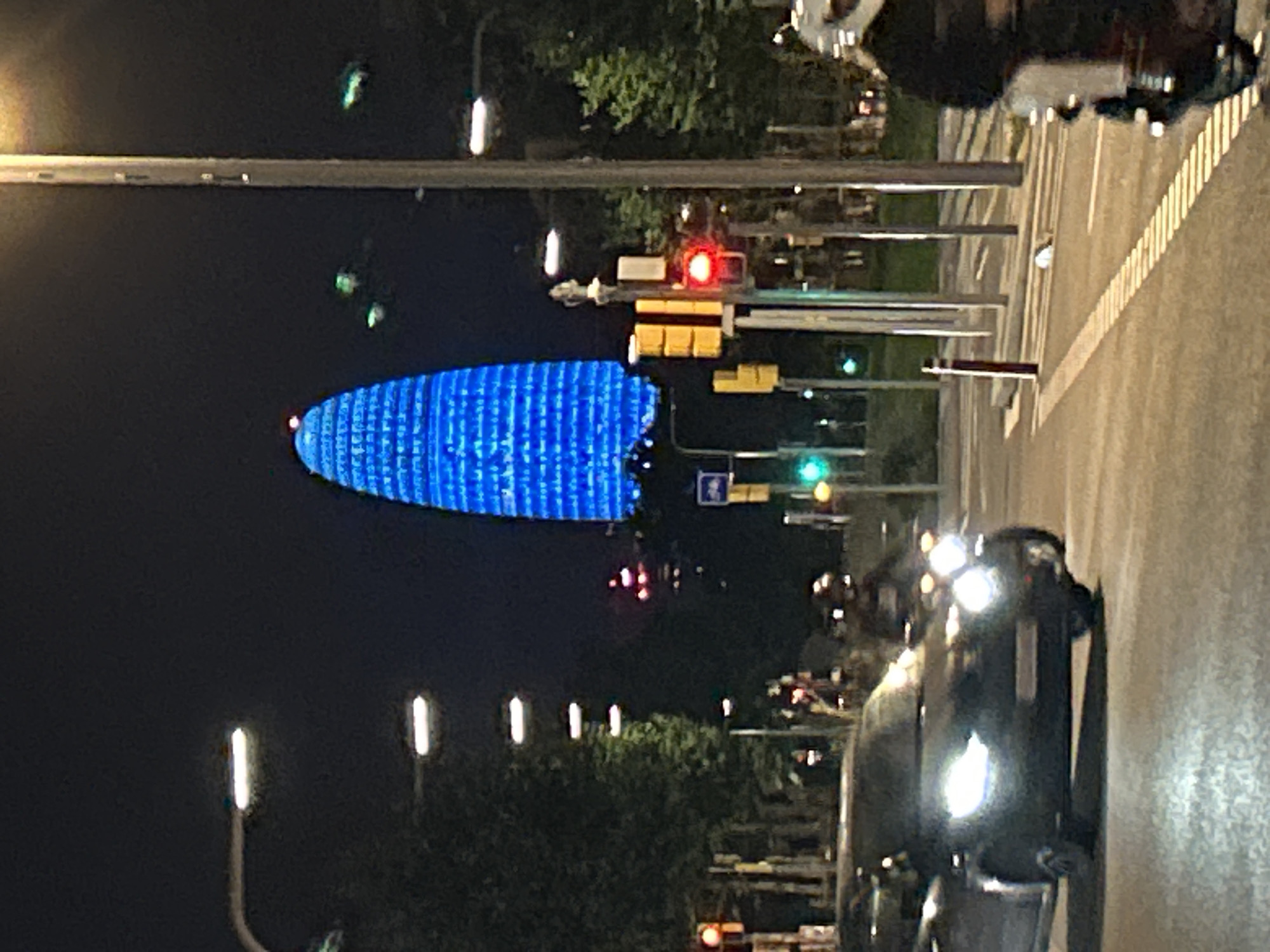 The image shows a street scene at night in Barcelona, Spain, with the iconic Torre Glòries building prominently lit in blue in the background. A black car with its headlights on is in the foreground, driving on a street. To the right, a person on a motorcycle, wearing a white helmet and a dark jacket, is visible. Traffic lights, streetlights, and trees line the street. The overall scene depicts a city at night with active traffic.