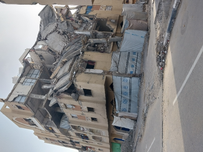 The image displays a severely damaged multi-story building in an urban setting, identified as Khan Yunis, Palestinian Territory. The building, likely a residential structure, exhibits extensive destruction, with several upper and middle floors collapsed or partially disintegrated. Concrete slabs are fractured and hanging, exposing twisted rebar and internal structural elements. A significant amount of rubble and debris has accumulated at the base of the building, indicative of a major destructive event.
Along the ground floor of the damaged building, several sections are covered with light-colored tarpaulins or heavy sheets, secured to create makeshift enclosures. These coverings suggest an attempt to provide shelter or cover for existing spaces. Visible on multiple tarpaulins are blue logos and text. The UNICEF logo, featuring the word "UNICEF" with a child icon, is clearly discernible on one of the coverings, indicating the use of humanitarian aid materials. Other blue labels with indistinct text, possibly in Arabic or English (e.g., "UNITED NATIONS"), are also present on the sheets.
Adjacent to the building is a paved street or road, marked by a white line. The edge of the road alongside the building is strewn with debris, including small rocks, dirt, and refuse, further highlighting the state of disarray. The scene is captured during daytime under a clear sky, with bright, even lighting. There are no people visible in the image. The overall impression is one of widespread devastation and an urgent need for humanitarian assistance.