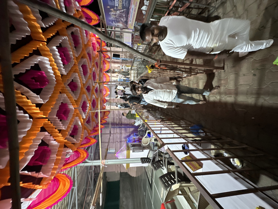 A festive scene unfolds with colorful decorations hanging overhead, predominantly in shades of orange, white, and pink. Below, a group of people are gathered, including men in white shirts and jeans. A metal railing runs along the side, and tables with white cloths and plates can be seen, hinting at a gathering or event. The lighting suggests it is evening or night.