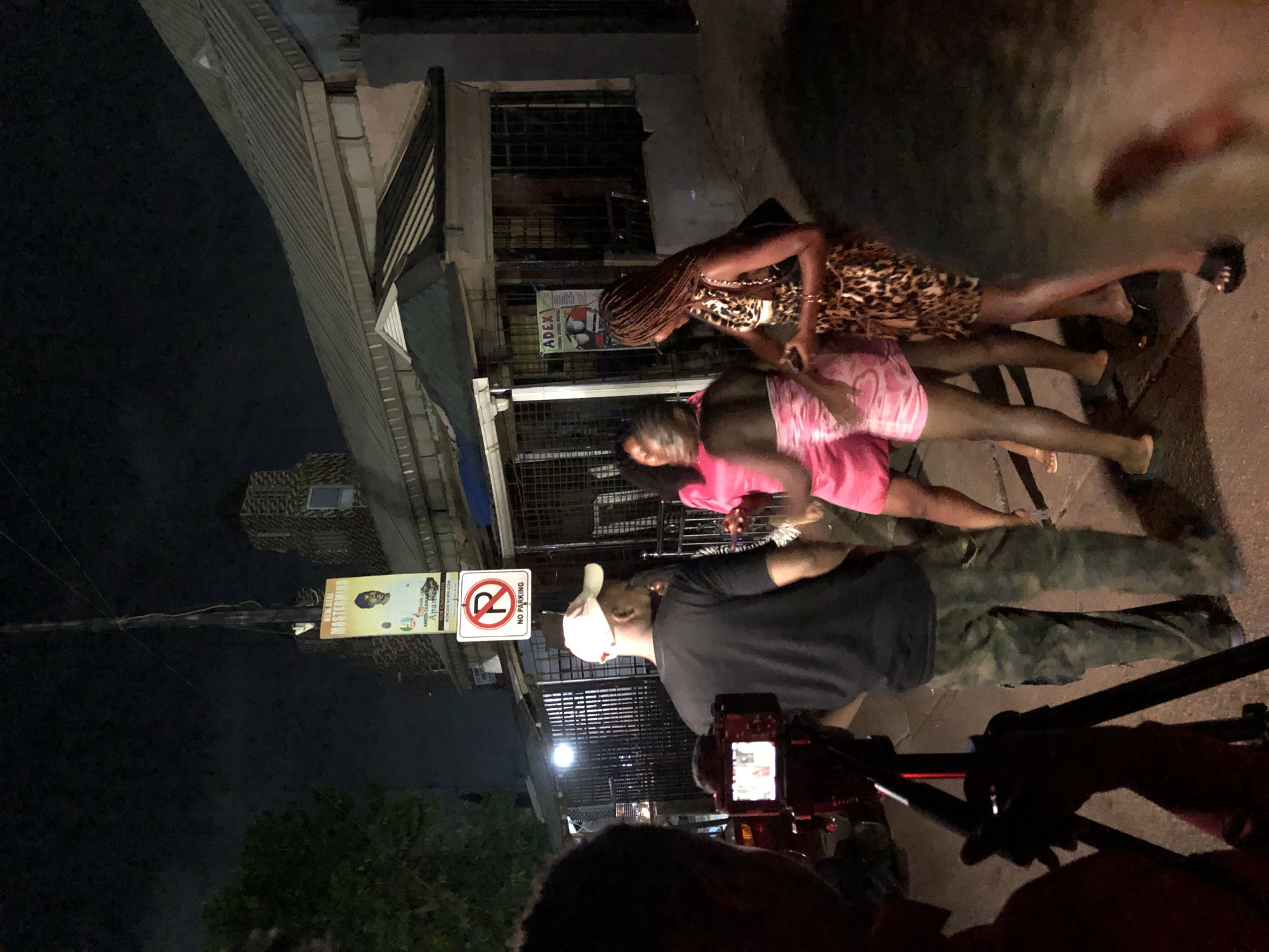 The image captures a night scene on what appears to be a street or alleyway in an urban setting. The sky is dark, and the area is lit by artificial light sources, possibly including those associated with a camera setup.
In the foreground, a person's head and shoulders are visible, operating a professional-looking camera mounted on a tripod or stabilizer. The camera's screen is illuminated. Facing the camera, slightly further back, is a man wearing a black t-shirt, camouflage patterned trousers, and a white baseball cap.
Two women stand beside him, positioned as if being filmed or photographed. One woman is wearing a leopard print dress and has braided hair. The other is dressed in a pink top and shorts. The women appear to be posing or standing in a casual manner for the camera.
The background consists of multi-story buildings with various architectural details, including slatted windows and security bars. One building features a banner advertising "APEX GYM & FITNESS CENTER". A street pole stands prominently, displaying a "NO PARKING" sign (red circle with a diagonal line over a 'P') and another banner. The second banner includes text like "NEW REAL MASTERPIECES" and what appears to be names or a brand, possibly "AMBO CLAVO" and "MORIOLA", next to a portrait of a man. The ground is a paved surface.
The overall activity suggests a media production, such as a video shoot or a photoshoot, taking place at night.