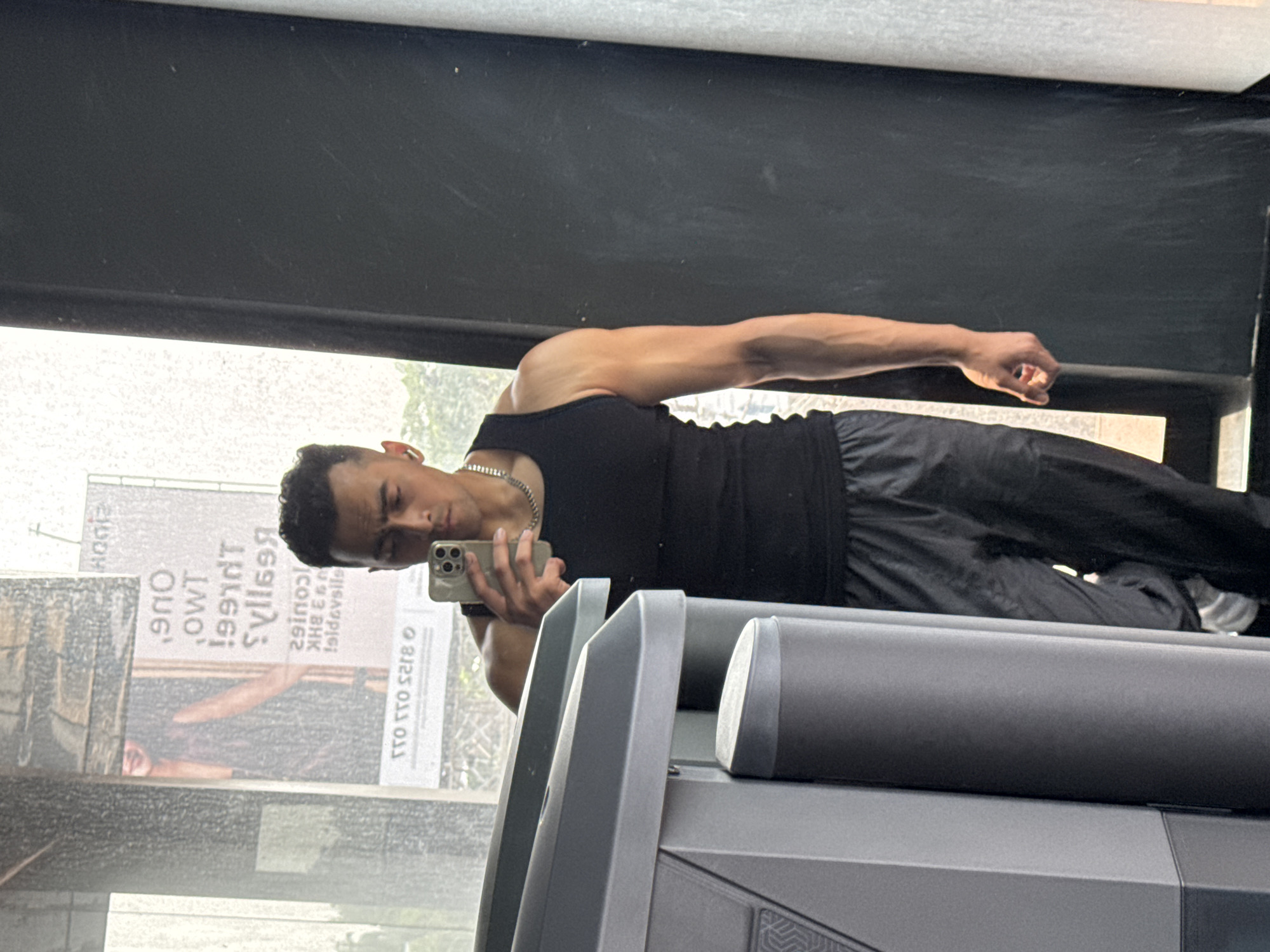 A muscular young man is captured in a selfie reflection, posing in what appears to be a gym setting. He is wearing a black tank top, dark athletic pants, and a silver chain around his neck. His left arm is extended and slightly bent, showcasing his bicep, while his right hand holds a light-colored smartphone (with multiple camera lenses visible) to take the picture. He looks directly towards the phone screen with a neutral expression.
The man is reflected in a large, clear mirror, which also reveals elements of the indoor gym environment, including a dark-colored piece of gym equipment, possibly a treadmill or similar machine, with gray accents and a textured pattern on its base.
Behind the man, through the mirror's reflection, an outdoor scene is visible, dominated by a large advertising billboard. The text on the billboard is reversed and slightly distorted due to the reflection. Visible text includes several words and numbers, indicative of a real estate advertisement: "HOME," "ONE," "TWO," "23 BHK" (likely referring to 2/3 Bedroom, Hall, Kitchen apartments), and sequences like "LCou162," "Syllae3," "l991dT," "XHHG," and "lsbdevals." A numerical sequence "8123 033 0TT" is also clearly visible, likely a contact number. The outdoor area appears bright, suggesting it is daytime. The overall scene suggests a moment of self-documentation within an urban gym environment in Kolkata, India.