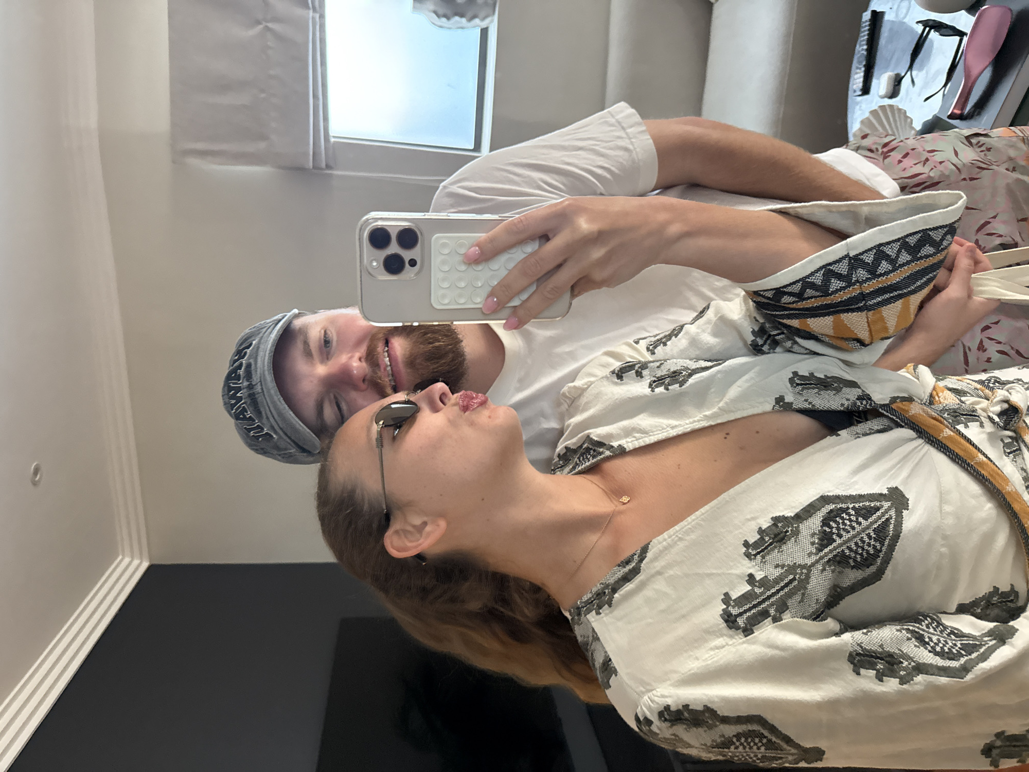 A man and a woman are posing for a selfie in what appears to be an indoor setting. The woman is in the foreground, wearing sunglasses and a white, patterned blouse. The man is behind her, wearing a grey baseball cap and a white t-shirt. He is holding a smartphone with a white pop socket. The background features a window and a couch. There is a patterned bag on top of the couch next to the seated person. There is no indication in the image that it was taken in Cape Town, South Africa.