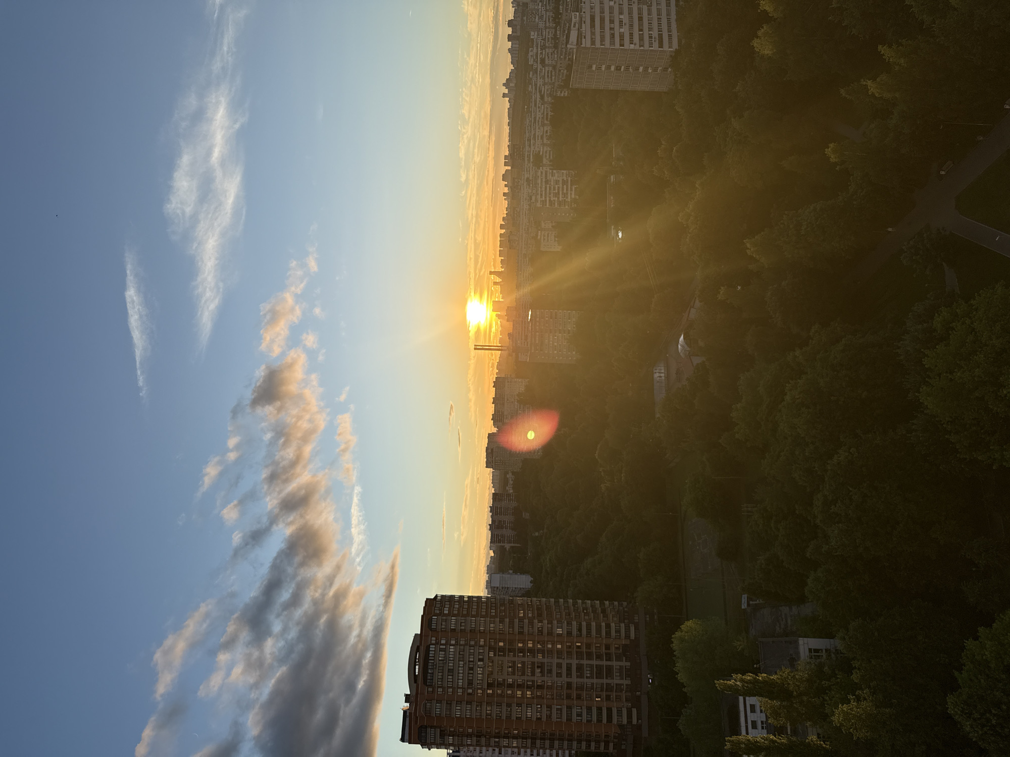 This is a high-angle, wide shot captured at sunset over a city that appears to be Moscow, Russia. The sky is a gradient of blue and orange, with scattered clouds tinged with the warm hues of the setting sun. The sun itself is a bright, slightly diffused orb near the horizon, casting long, golden rays across the scene.
In the foreground, to the left, is a tall, multi-story residential building, characterized by its red-brick facade and numerous windows. To the right and extending into the background is a dense canopy of green trees, suggesting a park or wooded area within the urban landscape. Beyond the trees, a panorama of city buildings, predominantly mid-rise residential blocks, stretches towards the horizon. Some of these buildings are lighter in color, contrasting with the darker trees and the sunset's glow.
The lighting indicates it is late afternoon or early evening. There are no discernible people or specific actions captured in the frame, focusing instead on the architecture, the natural elements, and the atmospheric effects of the sunset. The overall impression is one of an urban environment where nature and residential structures coexist, bathed in the dramatic light of dusk.