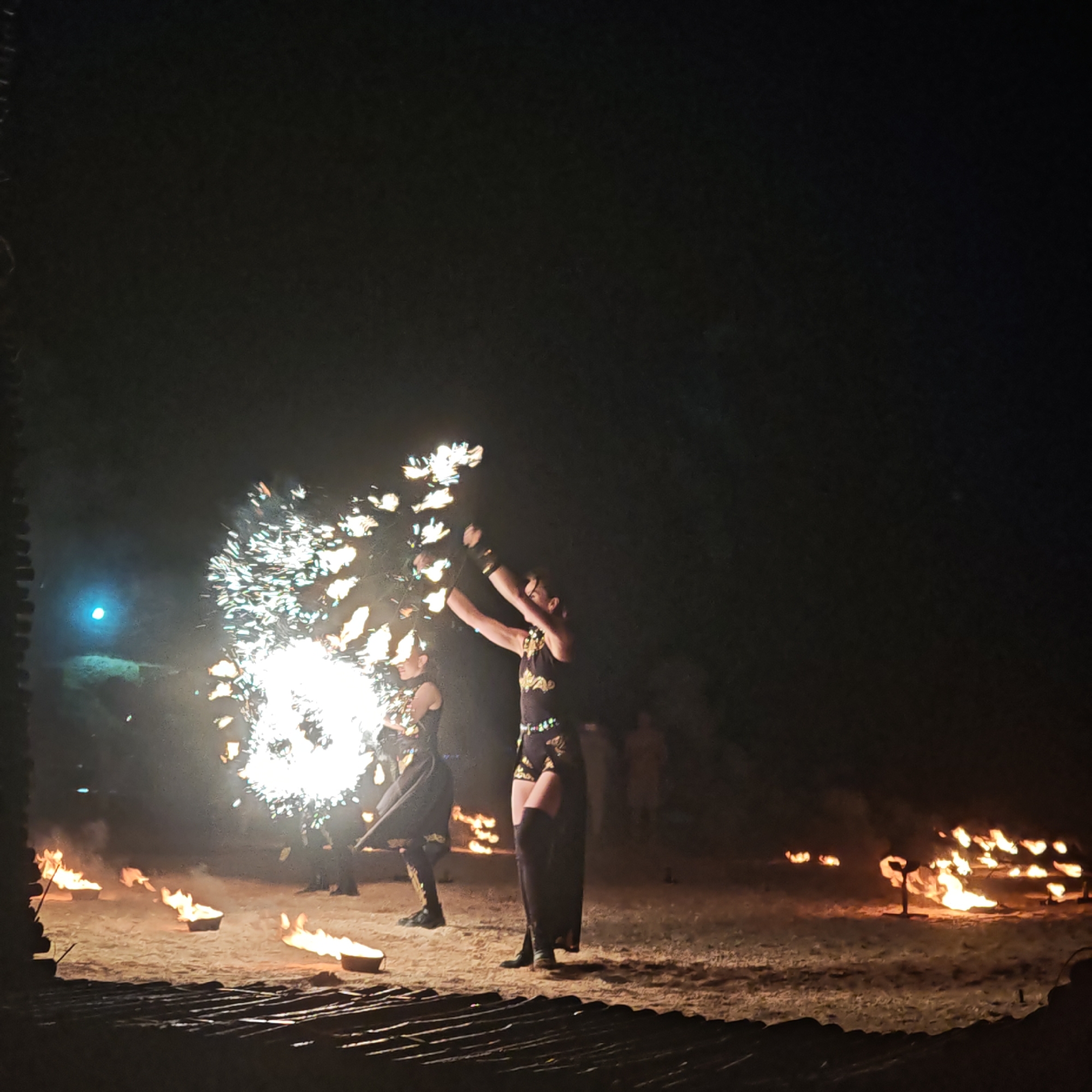 In the city of Shchelkino, Ukraine, a nighttime fire performance is underway on a sandy area, possibly a beach, with a dark, starless sky overhead. Two individuals are performing with fire. One performer, on the left, is wearing a dark costume with golden accents and is holding a fire prop, creating a large, sparkling burst of flames. Another performer, to the right, is also in a dark, ornate costume, possibly a skirt and high boots, and appears to be engaged in the fire display, with their arms raised. In the foreground, there are several small, lit fire bowls scattered on the sand. In the background, faint figures and trees are visible in the darkness. A bright blue light is present in the far left background, its source unclear. The overall scene depicts an energetic and visually striking fire show.