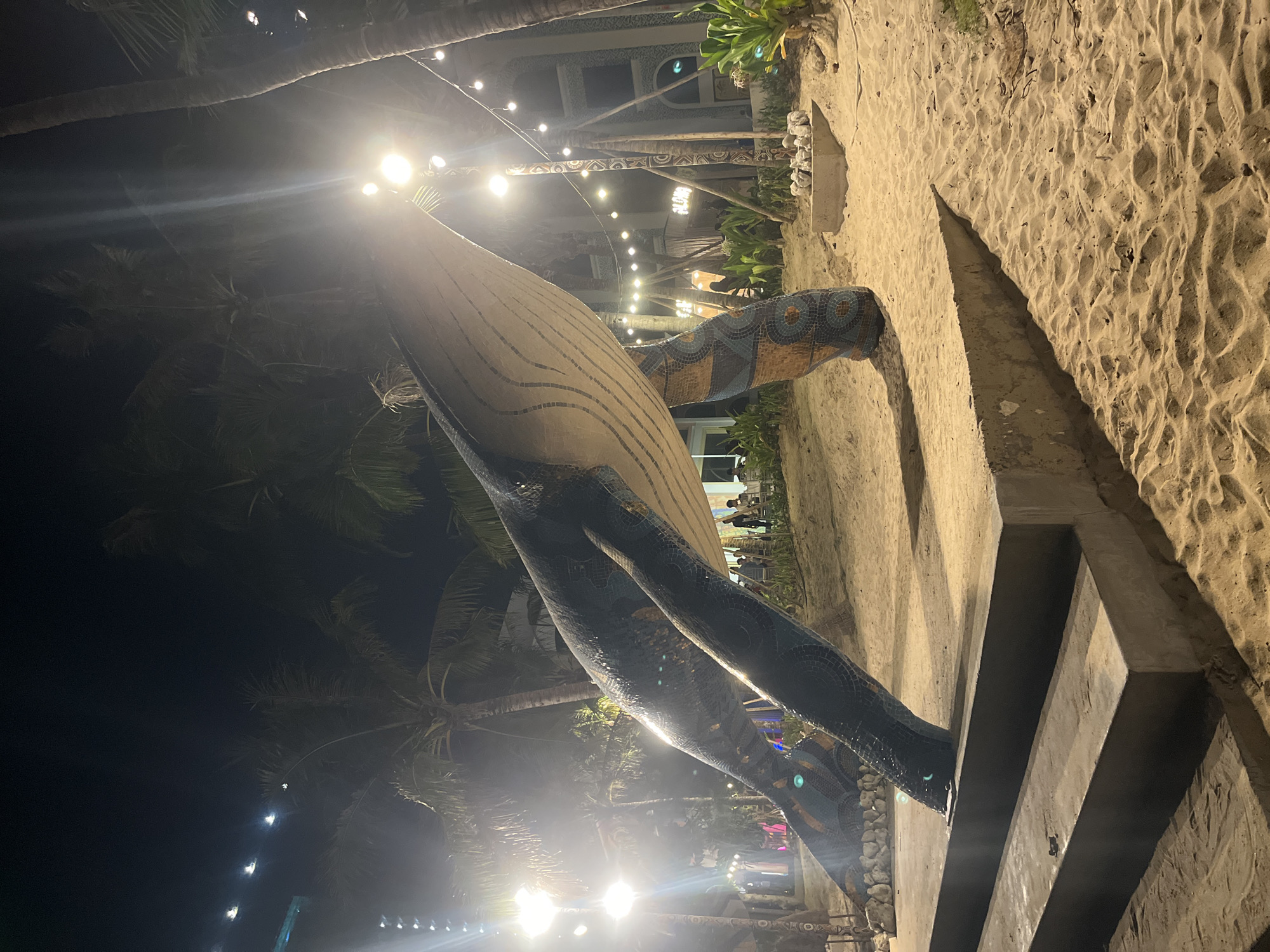 The image depicts an outdoor nighttime scene in Nha Trang, Vietnam, likely on a beach or a sandy area near a resort. The primary subject is a large, mosaic-covered sculpture of a whale, its body adorned with intricate patterns of blue, gold, and brown tiles. The sculpture is illuminated by bright lights, creating a dramatic effect against the dark sky.
The setting includes palm trees, evident by their fronds silhouetted against the light and the night sky. In the background, a building with architectural details and more lighting suggests a developed area, possibly a hotel or a venue. String lights are visible throughout the scene, adding to the festive and illuminated atmosphere.
The ground is covered in sand, with some concrete structures, including what appears to be a pathway or steps leading up to the sculpture. A few people are visible in the distance, moving around, suggesting some activity or event is taking place, though their specific actions are unclear. The overall mood is vibrant and visually striking due to the combination of the sculpture, the lighting, and the tropical environment. The time of day is clearly night, and the weather appears clear, allowing for the outdoor illumination to be effective. There is no visible text in the image.