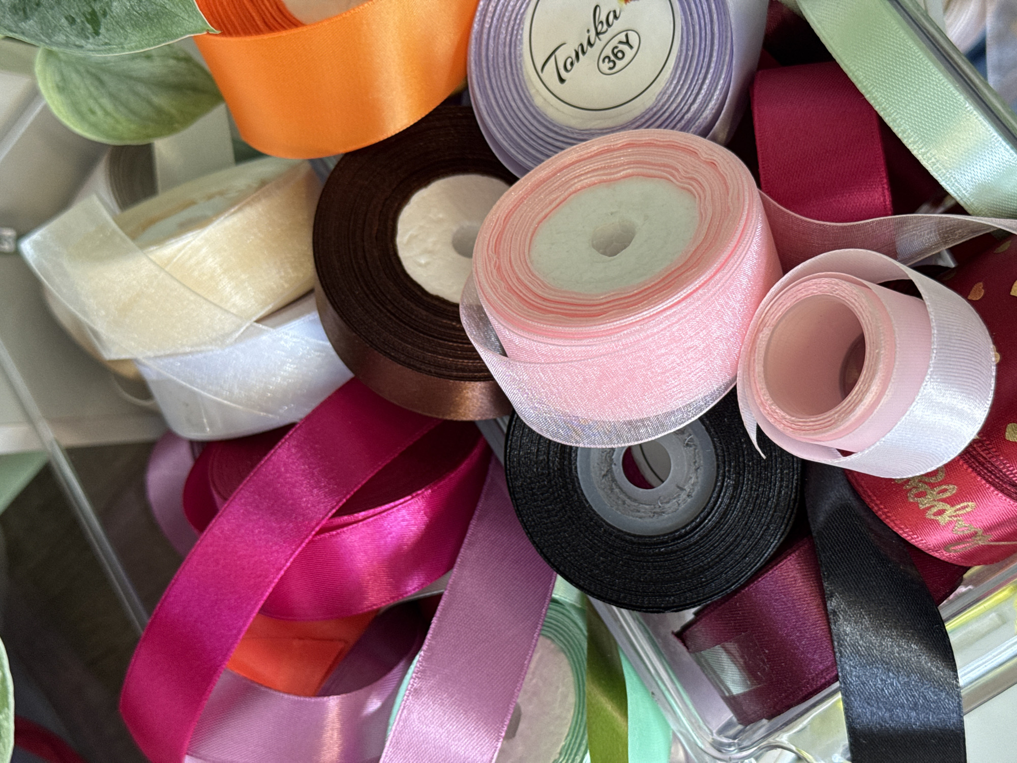 A close-up, high-angle shot displays a collection of colorful ribbon rolls and strands contained within a clear plastic bin. The ribbons are a vibrant assortment of pinks, purples, oranges, browns, blacks, reds, and greens, with some rolls featuring a shimmering or satin finish. The ribbons are neatly wound onto plastic spools, with some unfurled lengths spilling over the edges of the bin. The lighting is bright and even, highlighting the textures and colors of the materials. The background is softly blurred, suggesting a shallow depth of field. The image is devoid of people or any specific location markers.