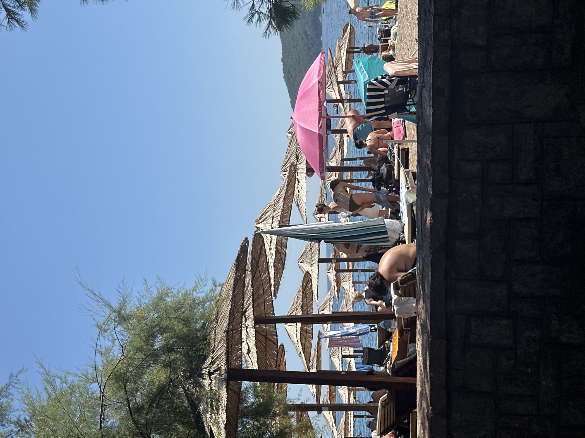 This is a medium-angle, daylight shot of a crowded beach in Budva, Montenegro. The beach features rows of thatched umbrellas casting shadows over loungers occupied by numerous people. The ocean is visible in the background, with a lush, green hill rising on the horizon. The foreground is dominated by a dark, stone wall.
In the foreground, a man with dark hair is sitting on a lounger, facing away from the camera. He is wearing dark shorts. To his left, another person is sitting in a deck chair, partially obscured.
Further into the scene, a bright pink umbrella stands out amongst the thatched ones. Several people are milling about, some in swimwear, preparing to relax or move along the beach. Towels and beach bags are scattered around. The atmosphere appears lively, indicative of a popular tourist destination.