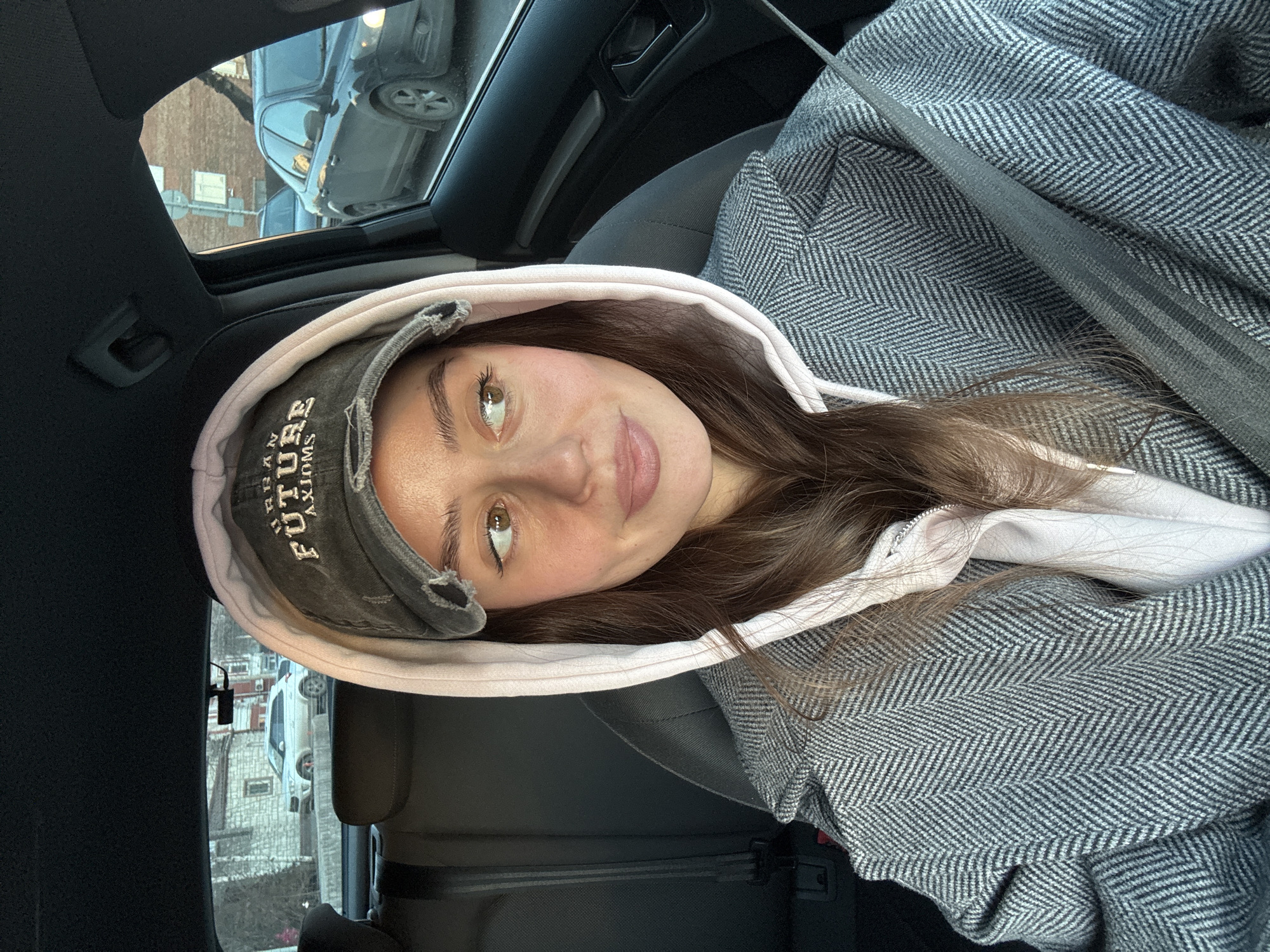 A woman with brown hair, wearing a backwards grey baseball cap with the words "Future Blooms" and a light pink hoodie underneath a herringbone-patterned grey coat, is sitting in the backseat of a car. The car appears to be in a city, with other vehicles visible through the windows and possibly a building. Based on the architectural style of the building visible through the window and general setting, this could plausibly be in the city of Krasnoyarsk, Russia. The woman is looking towards the camera with a slight smile.