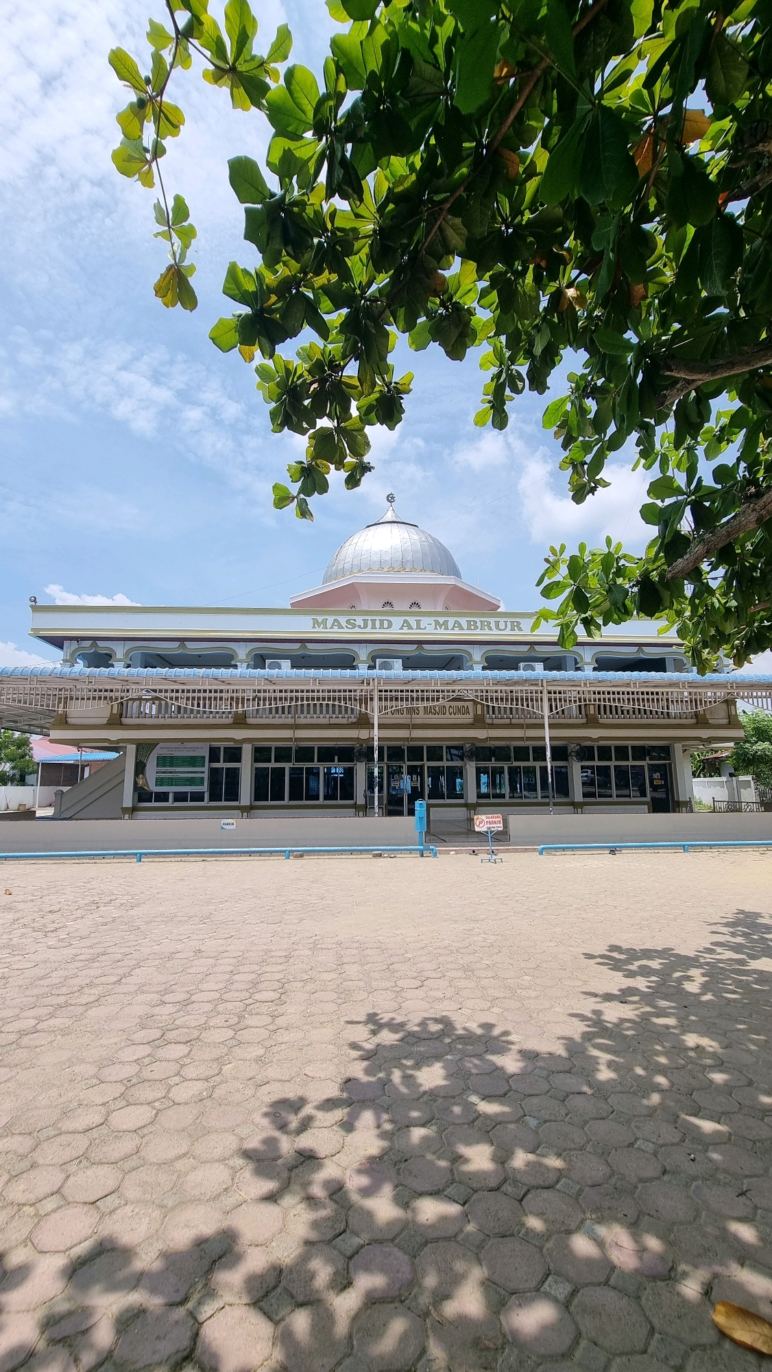This image depicts the Masjid Al-Mabrur in Lhokseumawe, Indonesia. The mosque features a prominent silver dome and is situated on a paved plaza. Trees with lush green leaves frame the top of the image, casting shadows on the plaza below. The sky is blue with scattered clouds. The facade of the mosque is light-colored with blue accents and numerous windows. Signs are visible on the building and on the plaza, one of which appears to be a "no parking" sign. No people are clearly visible in the frame. The overall scene suggests a sunny day in a public space.