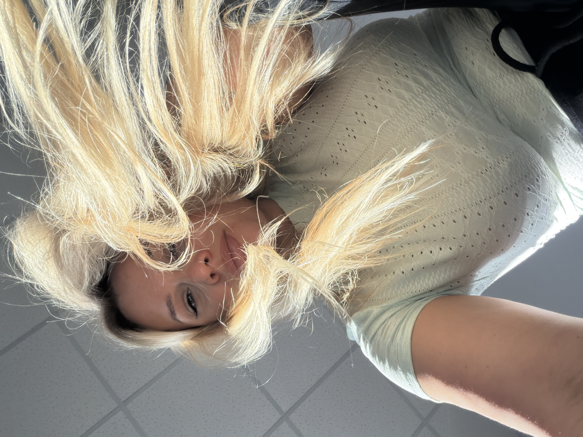 A low-angle photograph depicts an adult female looking downwards towards the camera. Her blonde hair is voluminous and appears windswept, framing and partially obscuring her face. Visible facial features include eyes, nose, and lips; one eye displays dark eyeliner. She is wearing a light green, short-sleeved top with a textured, perforated pattern. A portion of her arm is visible on the left side of the frame. The background consists of a light-colored suspended ceiling with a grid pattern, indicating an indoor environment. The specific location in Sarapul, Russia, cannot be determined from the visual information presented in the image.