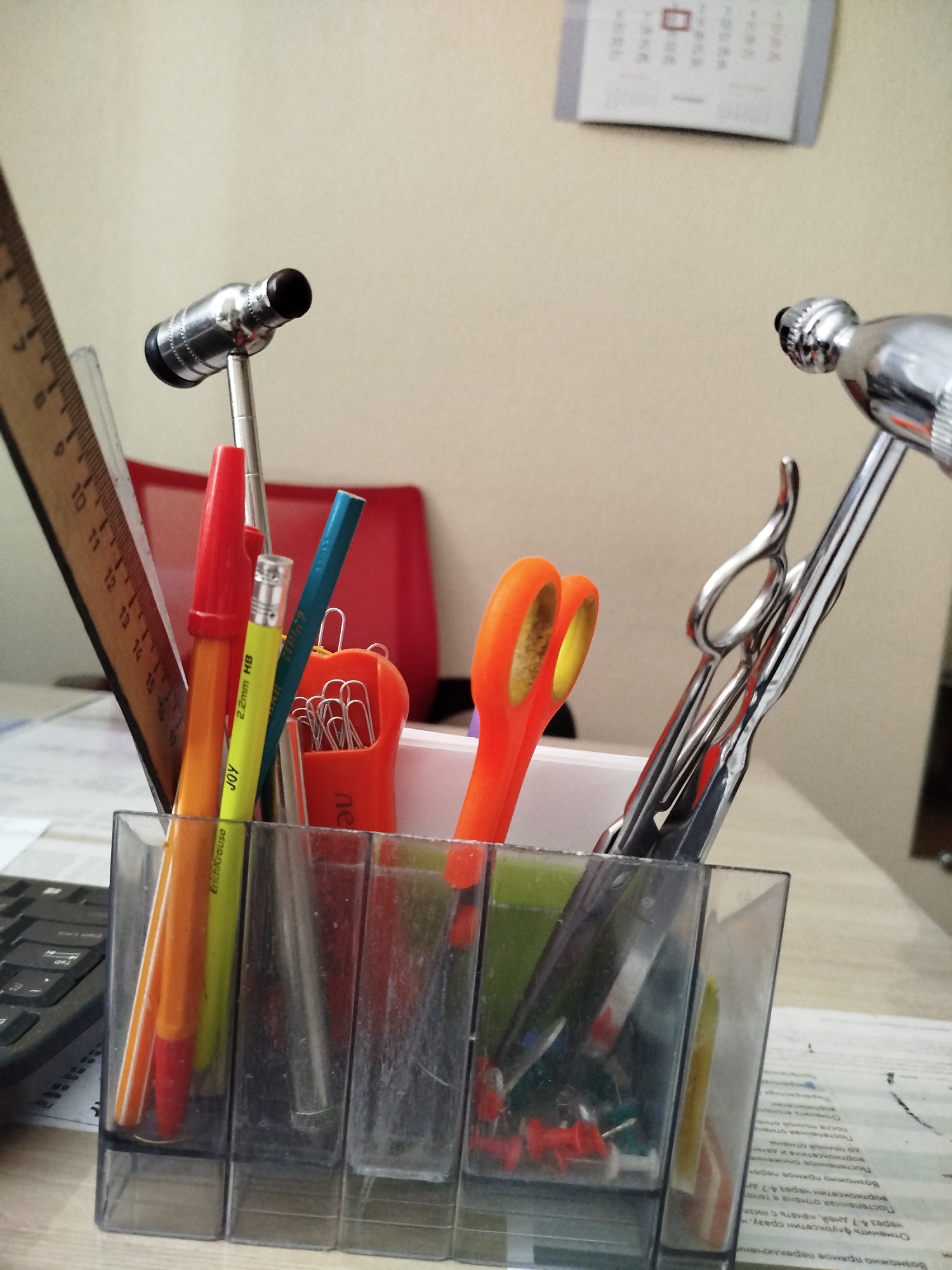 In Taganrog City District, Russia, a still life composition on a wooden desk features a clear plastic organizer filled with various office and medical supplies. The organizer contains a red marker, a yellow pencil, and several silver scissors. Outside the organizer, to the left, a wooden ruler with inch and centimeter markings is positioned vertically. To the right of the ruler, a silver reflex hammer with a black tip is visible. A second reflex hammer, also silver, is partially obscured on the far right. A red chair is visible in the background, and a calendar hangs on the wall above. The overall scene suggests a workspace or medical office.