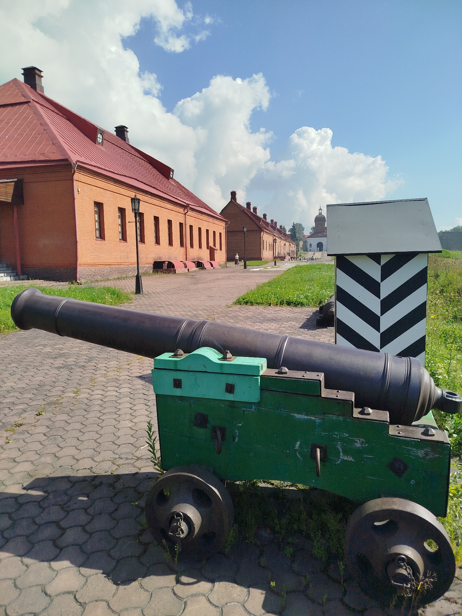 The image depicts a historical cannon mounted on a green wooden carriage, positioned on a paved courtyard. In the background, a long, red-brick building with a red roof stretches along the side of the courtyard, interspersed with lampposts. Further in the distance, a domed structure, likely a church or cathedral, is visible. To the right of the cannon, a black and white striped marker stands prominently. The scene is set under a bright blue sky dotted with fluffy white clouds. This image was taken at the Novokuznetsk Fortress in Novokuznetsk, Russia. There are no people actively engaged in any actions within the frame, and the overall environment suggests a historical site or museum. The lighting indicates it is daytime, likely with sunlight casting shadows on the paved ground.
