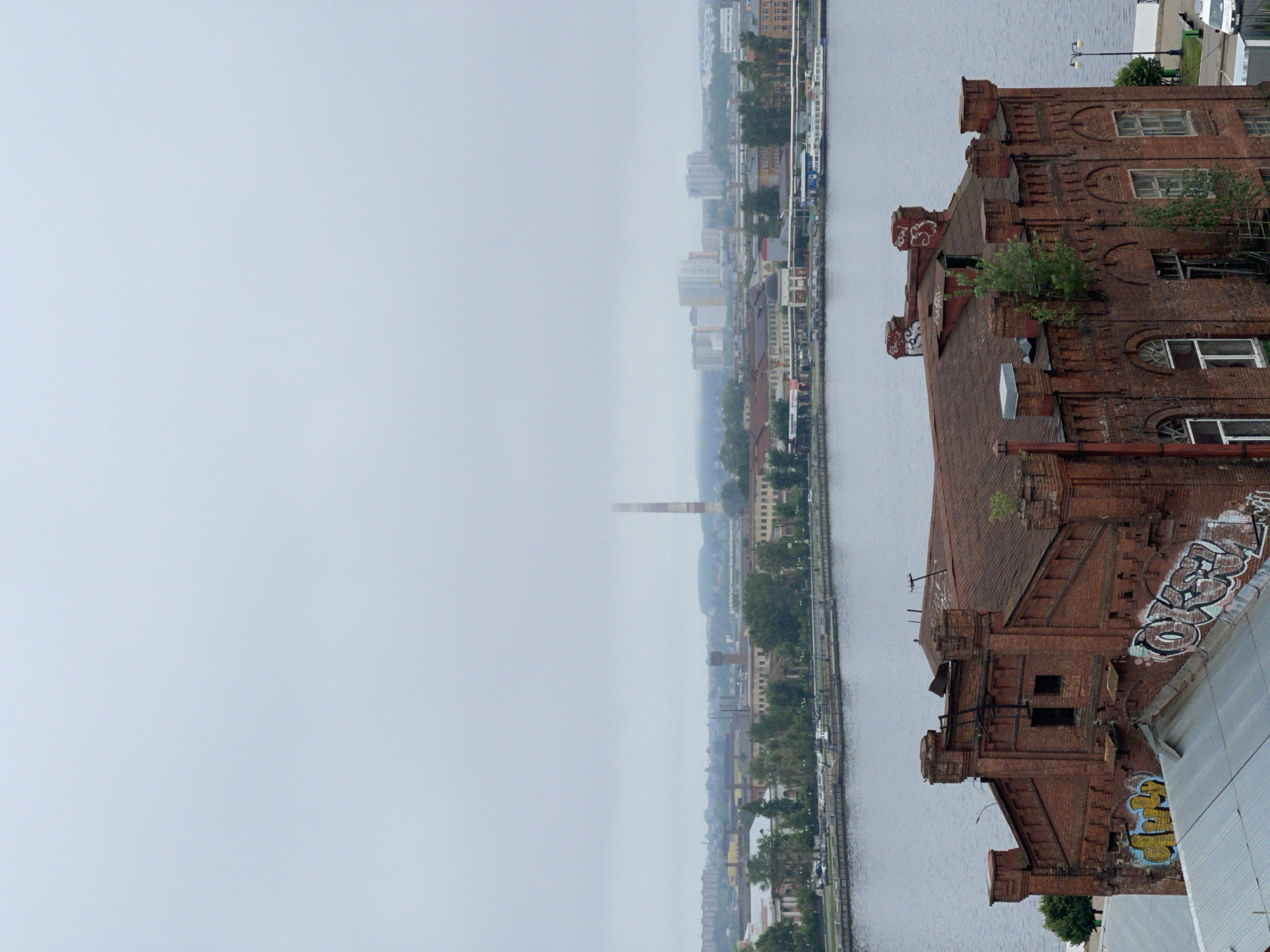 An aerial shot captures a brick building with graffiti and a large body of water in the foreground. Beyond the water, a cityscape unfolds with industrial buildings, a tall smokestack, and modern high-rise structures under a hazy, overcast sky. The brick building exhibits signs of disrepair, with weathered surfaces and vegetation growing on its facade. The graffiti on the building adds a layer of urban decay to the scene. The water appears calm, reflecting the muted tones of the sky and surrounding environment. The overall impression is one of an industrial port area with a mix of old and new architecture.