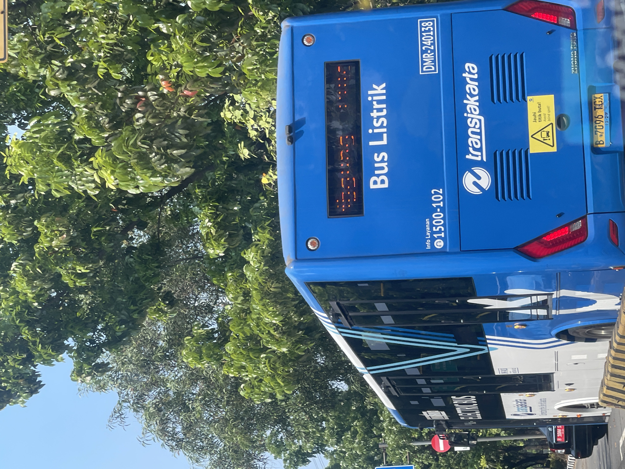 The image shows the rear of a bright blue Transjakarta electric bus in what appears to be East Jakarta, Indonesia, on a sunny day. The bus displays "Bus Listrik" (Electric Bus) on its back. Above this text, a digital display shows route information, though it is partially obscured and not fully legible. To the left of the bus, part of another vehicle and a traffic light are visible. The bus has the Transjakarta logo and branding. A yellow warning sticker with a graphic of a truck and text that reads "Jauhi titik buta! Awas blind spot!" (Stay away from blind spots! Beware of blind spots!) is affixed to the rear. The bus's license plate is "B 7096 TGX". Lush green trees with some red fruits are visible in the background, suggesting a street lined with vegetation.