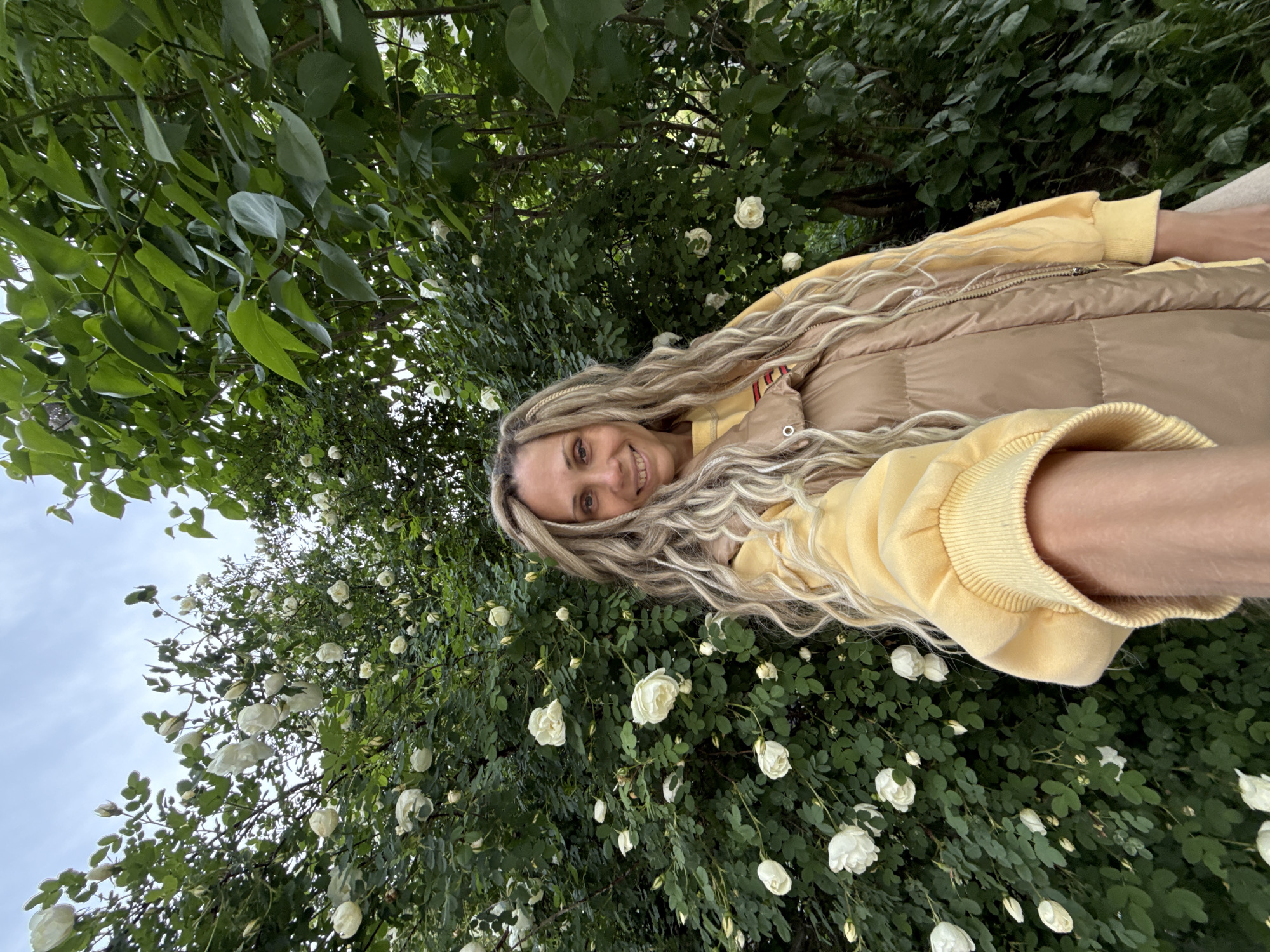 A woman is taking a selfie. She is in Rusanovo, Russia, surrounded by white roses and greenery. The woman has long, blonde hair and is wearing a yellow sweatshirt and a light brown vest. The background is a mix of green foliage and white rose blossoms. The sky is overcast.