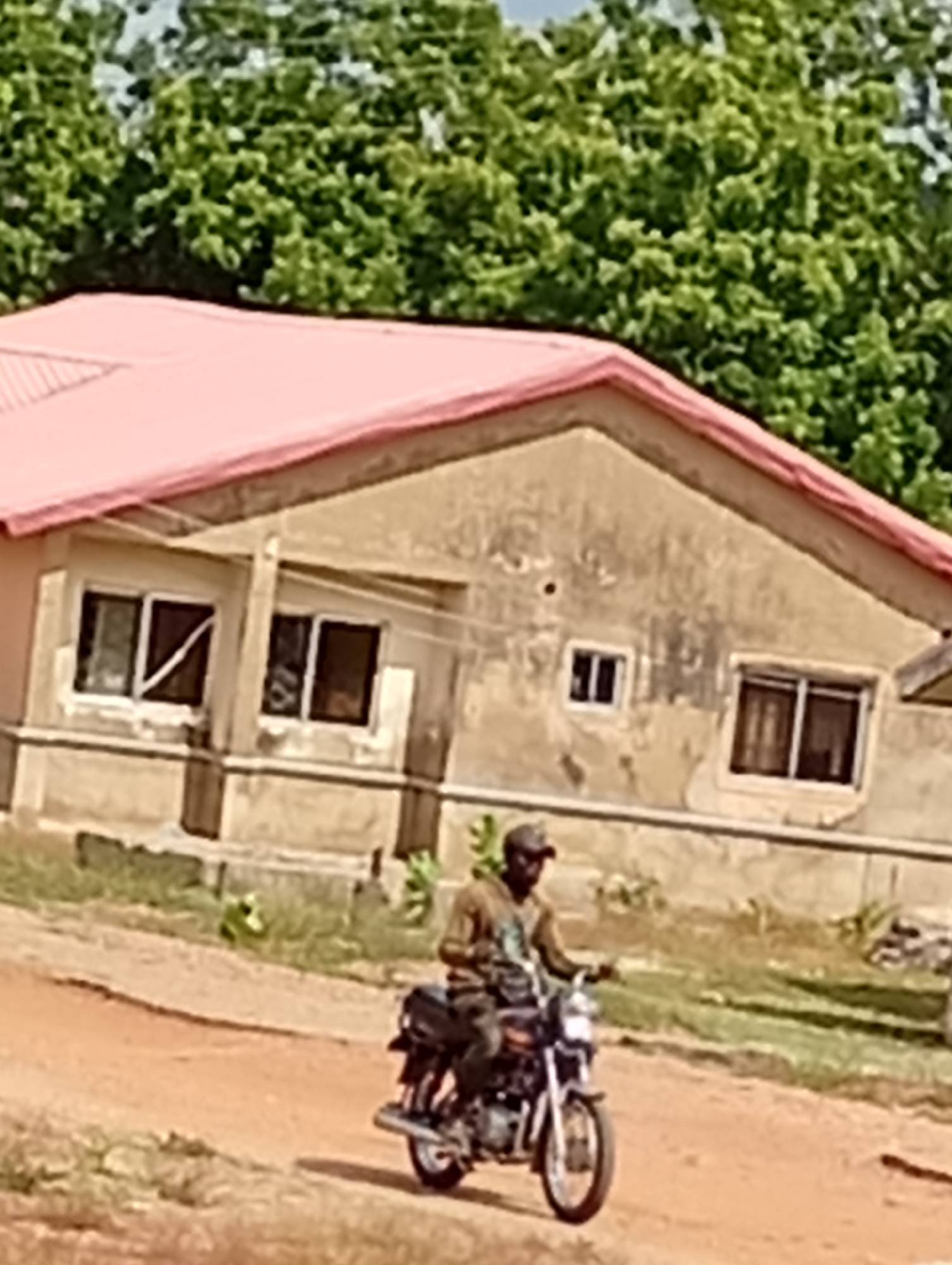 A man rides a motorcycle on a dirt road in front of a single-story building with a pink roof and multiple windows. The scene appears to be located in an unidentifiable area of the city of Unknown, Unknown. The man is wearing a cap and clothing that is unclear due to the image's resolution. Trees are visible in the background.
