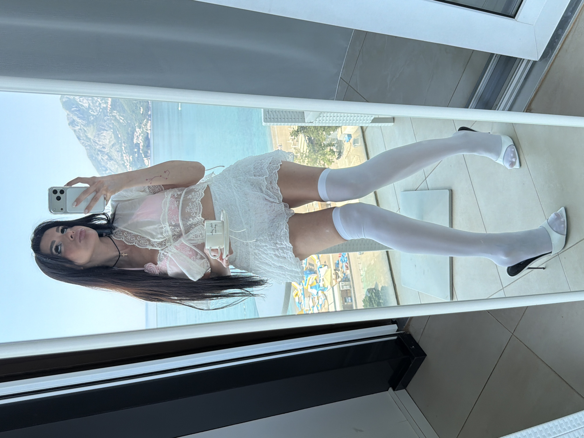 A woman is taking a selfie in a mirror. She is wearing a pink top with a white lace negligee, white knee-high socks, and white heels. Her hair is long and dark. The background visible through the mirror shows a beach resort in Boreti, Montenegro. The shot includes the sea, surrounding mountains, and some of the buildings within the resort. The floor beneath her feet is tiled. A black and white table is also in view.