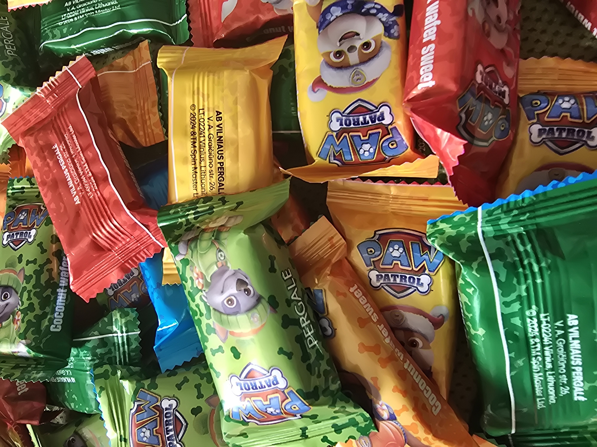 A close-up, cluttered view shows an assortment of individually wrapped candies, mostly in red, yellow, and green packaging. Many of the packages feature the branding "PAW PATROL" with character images from the show. Inscription visible on the packages indicates "AB VILNIAUS PERGALE, V. A. Graičiūno str. 26, LT-02241 Vilnius, Lithuania, ©2024 & TM Spin Master Ltd." This points to the location of Vilnius, Lithuania, and the manufacturer is AB VILNIAUS PERGALĖ. Flavors such as "Coconut wafer sweet" are also indicated on the packaging. The candies are piled together.