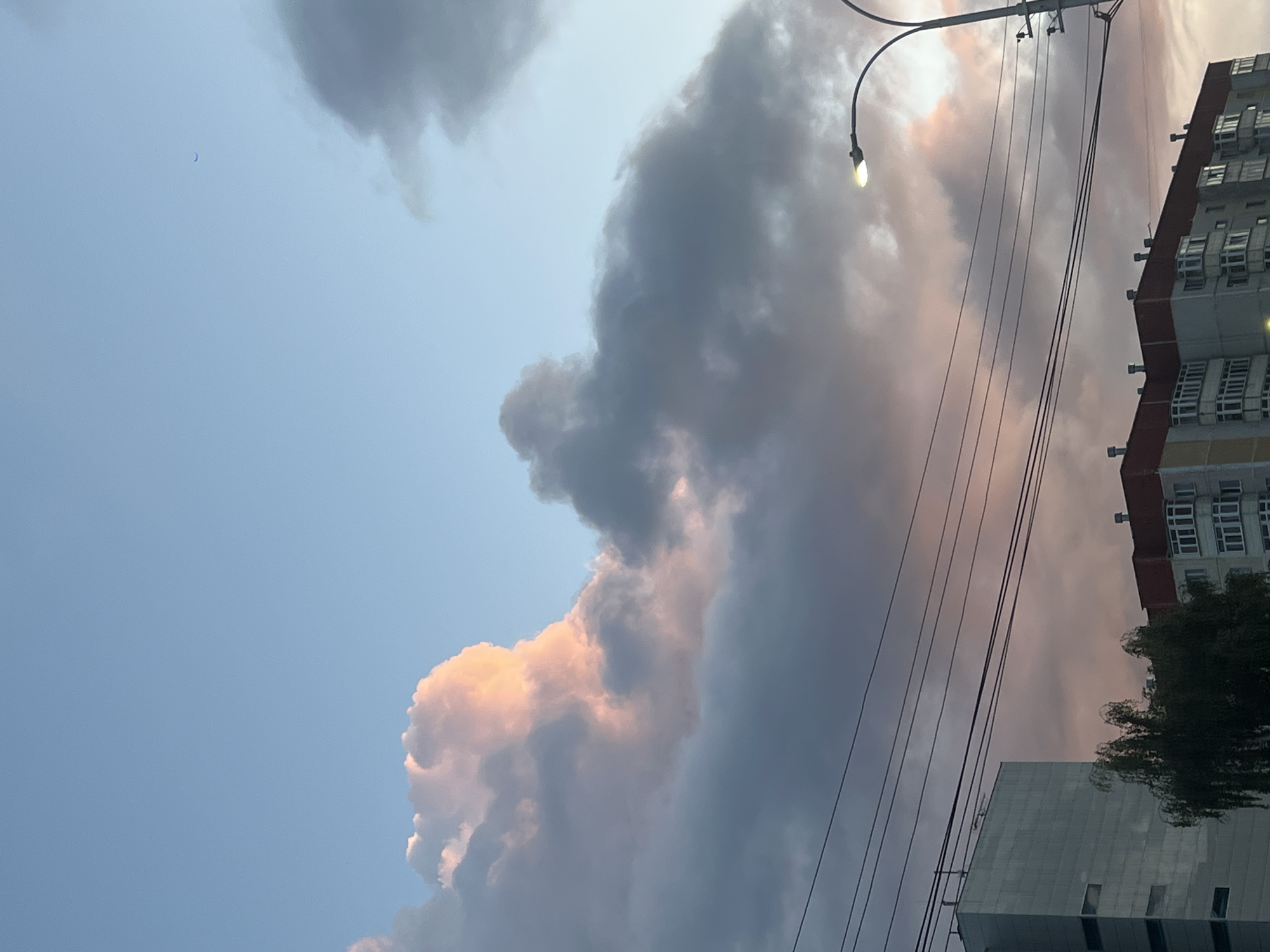 A low-angle, medium shot captures a cloudy sunset sky over Surgut, Russia. The sky is a gradient of light blue at the top, transitioning to pink and gray clouds below. A faint crescent moon is visible in the upper right quadrant. In the foreground, power lines stretch horizontally across the frame. Below the power lines, the tops of two multi-story buildings and a tree are visible. One building is tall and modern, with a gray facade and rectangular windows. The other building is shorter, with a red roof and many windows. A streetlamp with its light on is situated to the right of the frame. The overall impression is a typical urban evening scene.