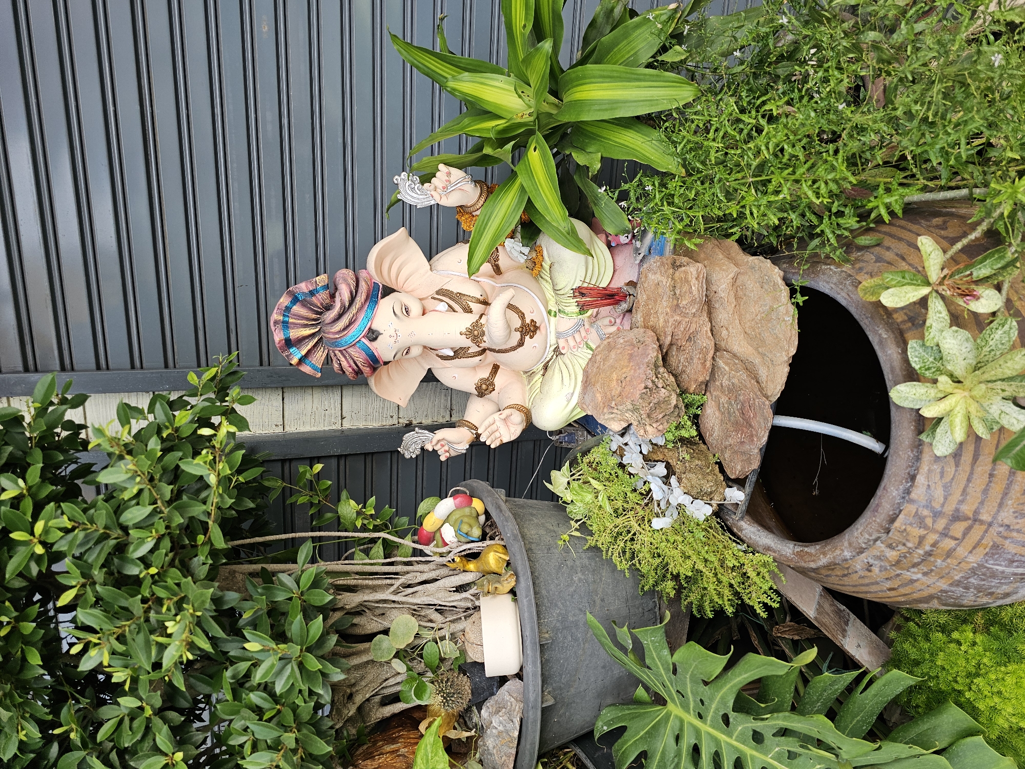 A statue of the Hindu deity Ganesha, adorned with a colorful turban, sits in a garden setting. The statue is light pink in color and is seated with its hands raised. In front of the statue, a small, rocky waterfall flows into a large, brown ceramic pot. Several green plants surround the statue and the waterfall, including a large-leafed monstera plant and a tall, thin-leafed plant with yellow and green stripes. To the left of the statue, a large potted tree with exposed roots is visible. Various small figurines, including what appears to be a frog and a pair of yellow animal figures, are placed in the potted tree. In the background, a dark gray, corrugated metal door is visible. The scene appears to be outdoors in an urban environment, possibly a residential area or a shop in Bangkok, Thailand, characterized by the tropical plants and the common use of such rolling metal doors.