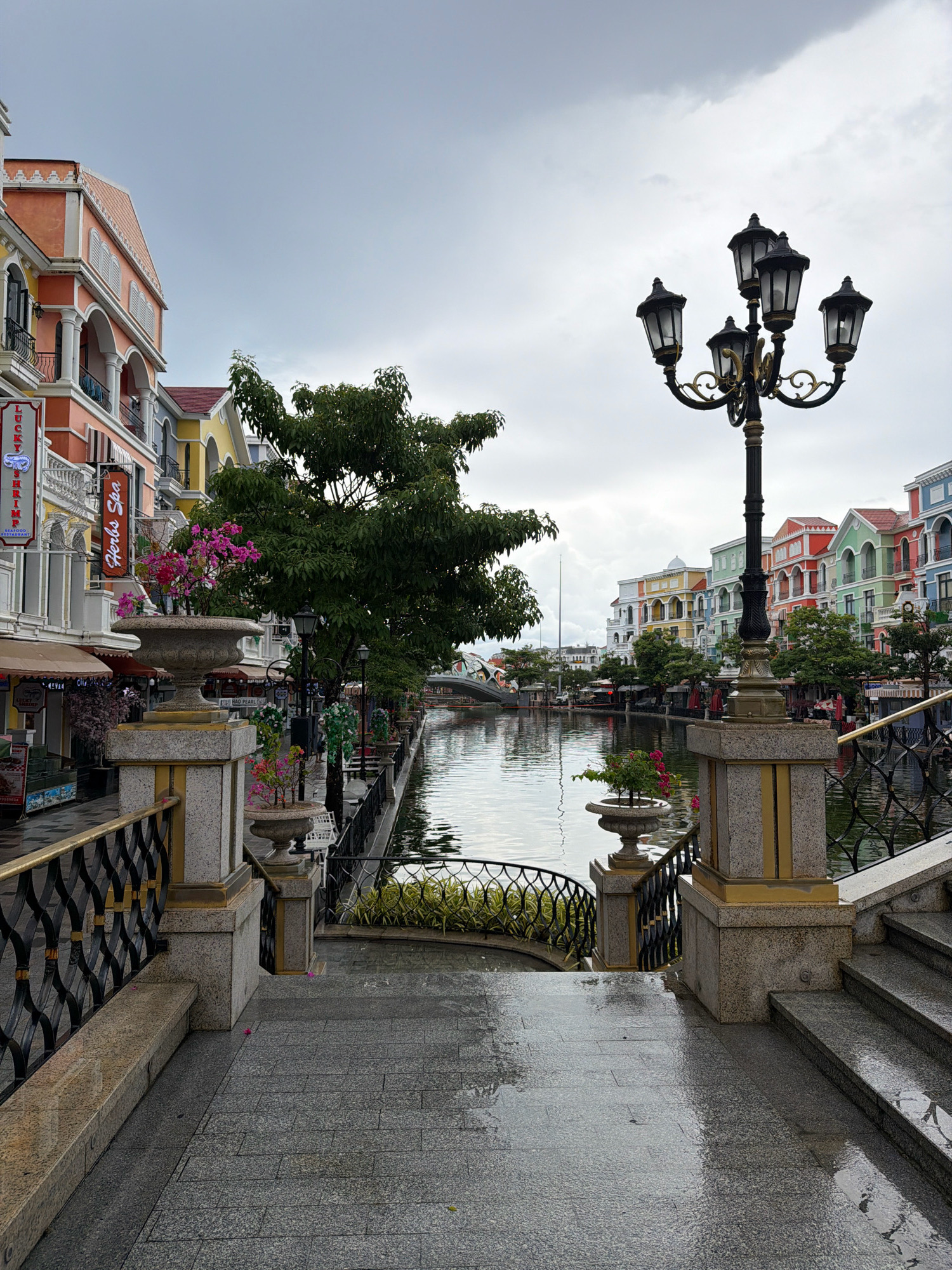The image depicts a picturesque canal-side promenade in Phu Quoc, Vietnam, likely within the Sun Premier Village Primavera complex. The architecture is vibrant and European-inspired, with pastel-colored buildings featuring arched windows and balconies lining the waterway. A large, ornate multi-light lamppost stands prominently on the right side of the frame, its dark metal structure contrasting with the bright buildings and overcast sky.
The canal itself is calm and reflective, with a dark, still surface. Lush green trees and manicured plants line the banks, adding a touch of nature to the urban landscape. On the left side of the promenade, a large stone planter holds vibrant pink flowers, while smaller planters with greenery are visible along the canal's edge. The ground is paved with grey granite tiles, which are wet and reflective, suggesting recent rain. A decorative black metal railing runs along the promenade, separating it from the canal. Stairs leading down to the water's edge are visible on the right. The overall atmosphere is serene and somewhat subdued, possibly due to the overcast weather. There are no people visible in the frame, and no specific actions are taking place. The scene is static, focusing on the architectural beauty and tranquil environment of the location.