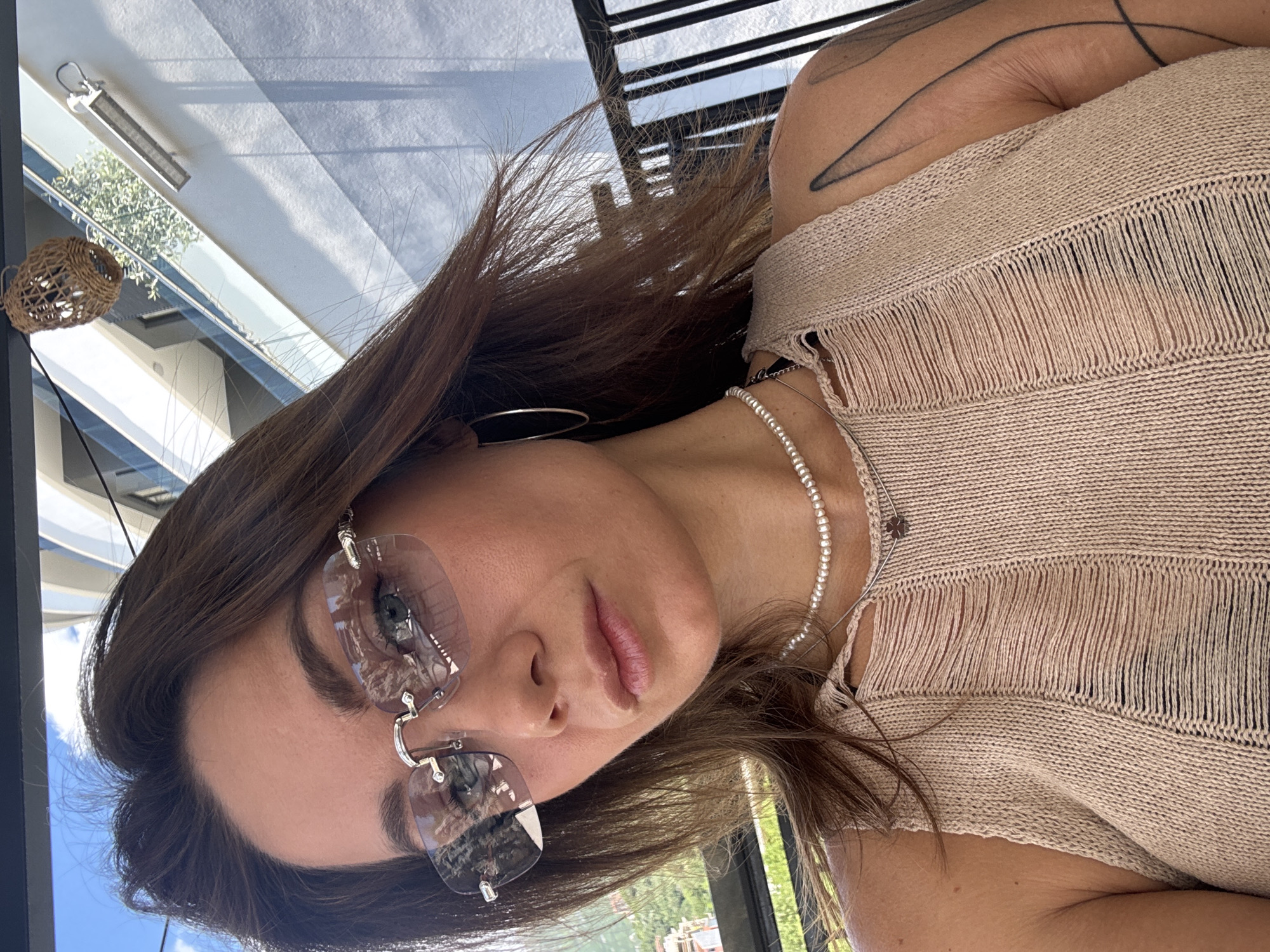 A woman is posing in front of a building in Boreti, Montenegro. She is wearing sunglasses, a necklace, and a tan, fringed top. Her hair is brown, and she has a tattoo on her shoulder. The background includes parts of building structures.