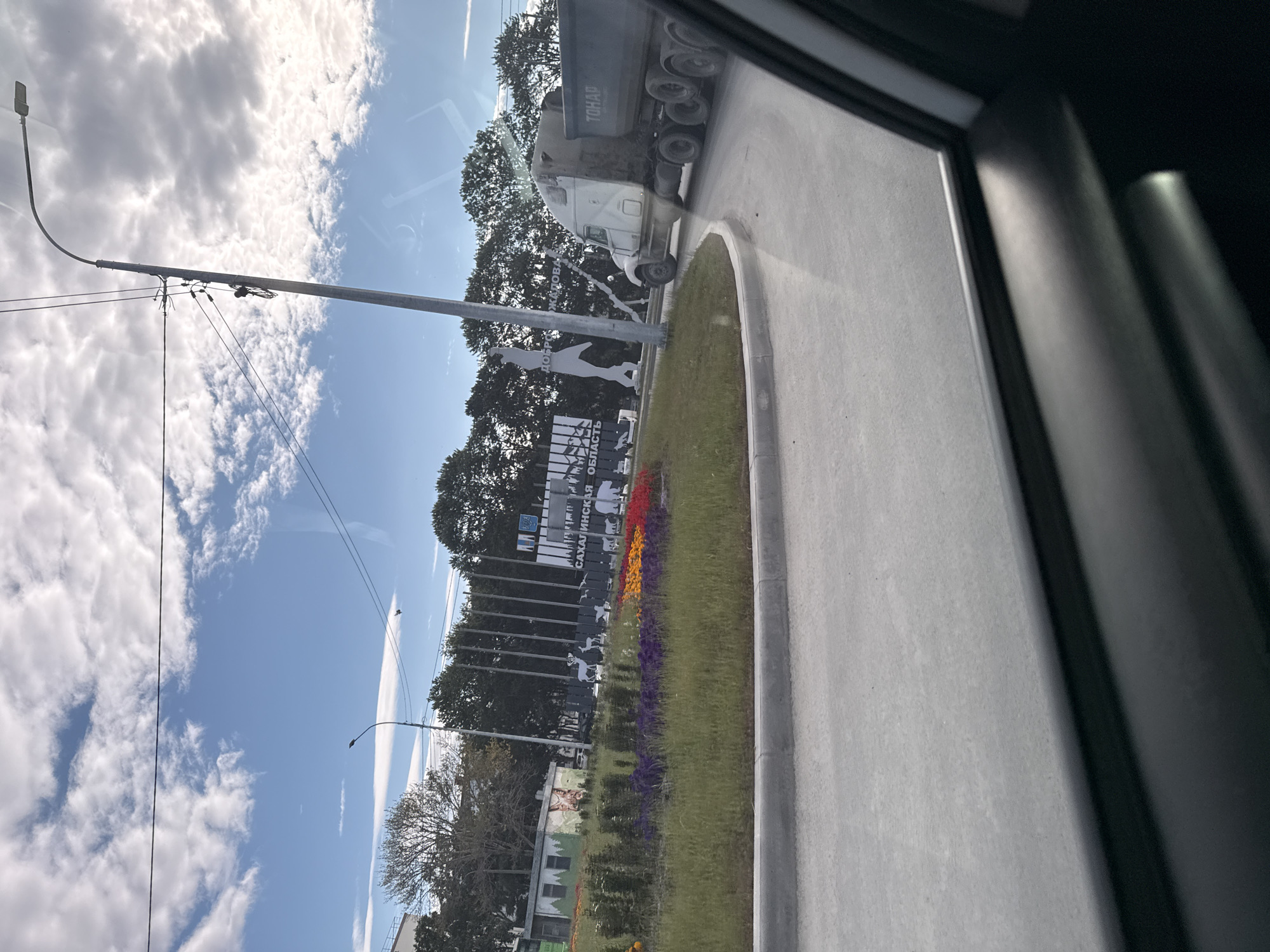 A white semi-truck with a dark gray dump trailer is turning a corner on a paved road in Yuzhno-Sakhalinsk, Russia. The truck's trailer is labeled "ТОНАР". In the background, there is a circular green island with a vibrant flowerbed of red, orange, and purple flowers. Behind the flowerbed, there is a monument with silhouetted animals (a deer, birds, and a bear) and text that reads "ДОБРО ПОЖАЛОВАТЬ" and "САХАЛИНСКАЯ ОБЛАСТЬ". To the left of the monument is a small, white building with graffiti on its side. The sky is blue with scattered white clouds. A street lamp is visible on the right side of the image. The photo is taken from inside a vehicle, as indicated by the car interior visible in the foreground.