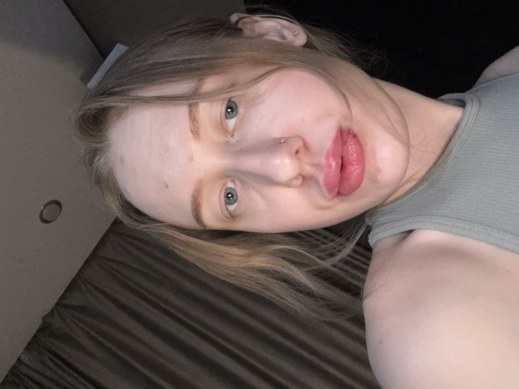 A close-up portrait depicts a young woman with fair skin, light brown hair, and blue eyes. She has a nose piercing and what appears to be a small earring. She is wearing a gray tank top. The background features a wall with a light switch and a bed or upholstered surface with dark brown stripes. No evidence suggests the location is Rostov-on-Don, Russia.