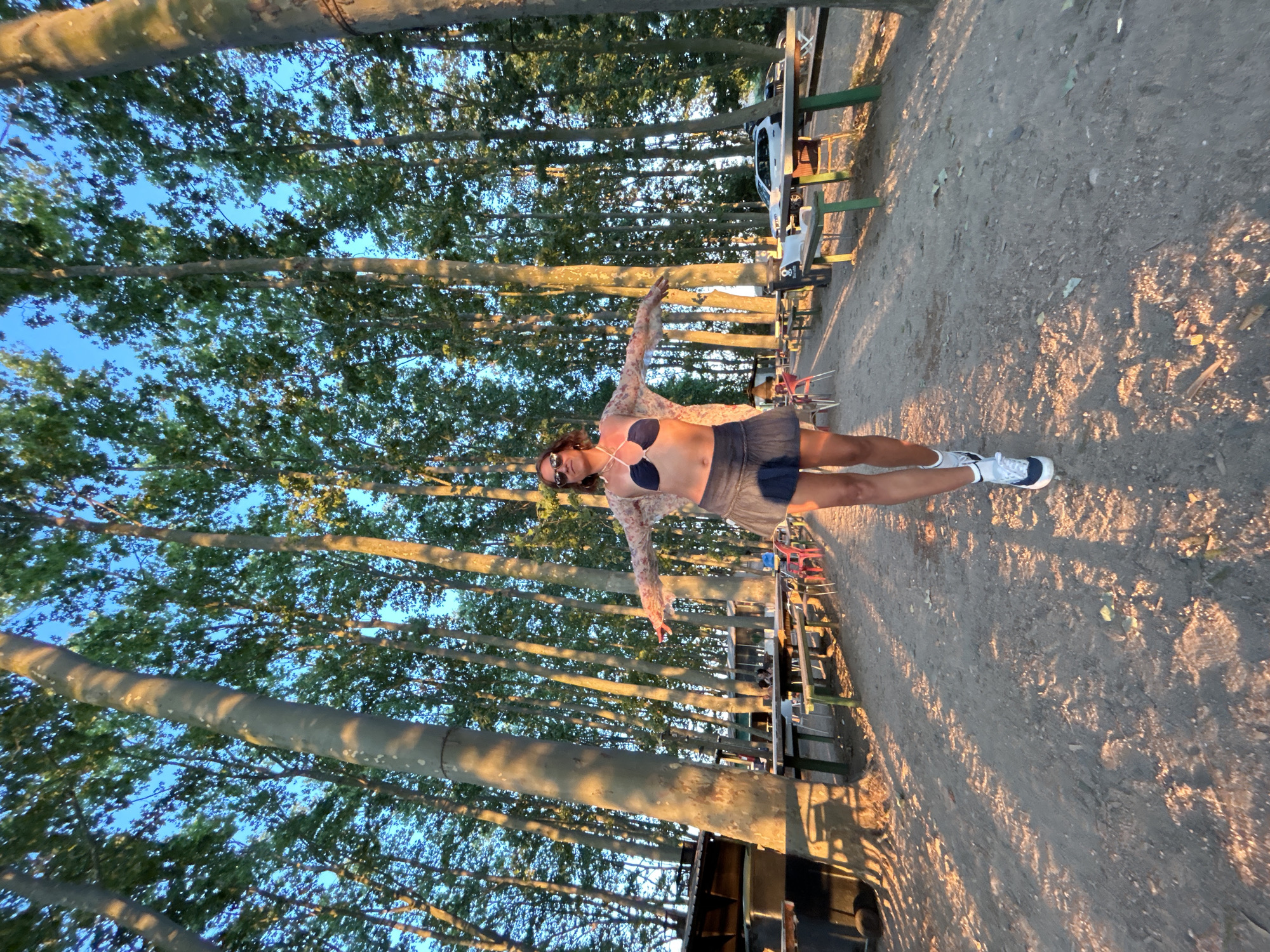 A full-shot, eye-level, outdoor photo captures a woman standing in the middle of a path lined with tall trees. She is wearing a dark blue bikini top, a dark blue skirt, a sheer, patterned top, white socks, and white sneakers. Her arms are outstretched to the sides. The path is dirt and gravel, with dappled sunlight and shadows from the trees. On either side of the path are picnic tables and benches, some with people seated at them. In the background, more trees and a white car are visible. The sky is blue and clear. This scene is located in Santa Cristina d'Aro, Spain.