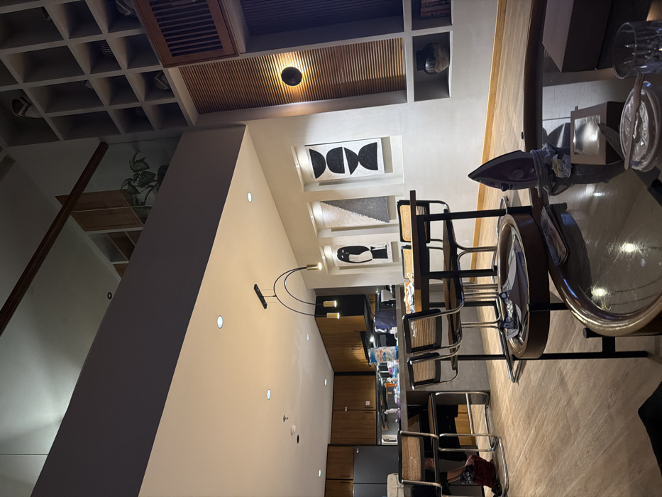 This image depicts a modern, multi-level residential interior located in Tibubeneng, Indonesia. The perspective is from a lower living area looking towards a kitchen and dining space, with a mezzanine visible above.
In the immediate foreground, a round, dark wood coffee table with a reflective glass top is present. On it, a purple clothes iron rests upright on a crumpled white cloth or paper, positioned slightly off-center. To the right of the iron, a clear glass and a white circular coaster are visible. A rectangular brown box and some papers or currency are also on the table.
The midground features a dining area. A dark rectangular dining table is surrounded by four chairs with dark metal frames and cane-woven backs and seats. Above the table, a contemporary linear pendant light fixture with two gold-colored cylindrical lights hangs from the ceiling. The wall behind the dining table displays three vertical framed abstract art pieces, primarily featuring black, white, and gray geometric or organic shapes. To the right, a built-in wall unit incorporates horizontal wooden slats, a circular wall sconce emitting warm light, and various open shelves containing decorative objects like vases and books.
In the background, a kitchen area is visible, characterized by light wood-toned upper and lower cabinets. A dark gray island counter extends into the room, with two bar stools matching the dining chairs situated beneath it. A large black refrigerator stands to the left. Above the island, a range hood is present, and various containers and bottles are visible on the countertop. A person is partially visible, seated on the left-most bar stool, with only their lower body and legs discernible.
The overall environment features light-colored walls and light-toned wood-look flooring throughout. The ceiling contains multiple recessed lights. Above the main living space, a mezzanine level is visible with a glass and wood railing. The mezzanine also shows built-in shelving and a green potted plant. The space is illuminated by artificial lighting, creating a warm ambiance.