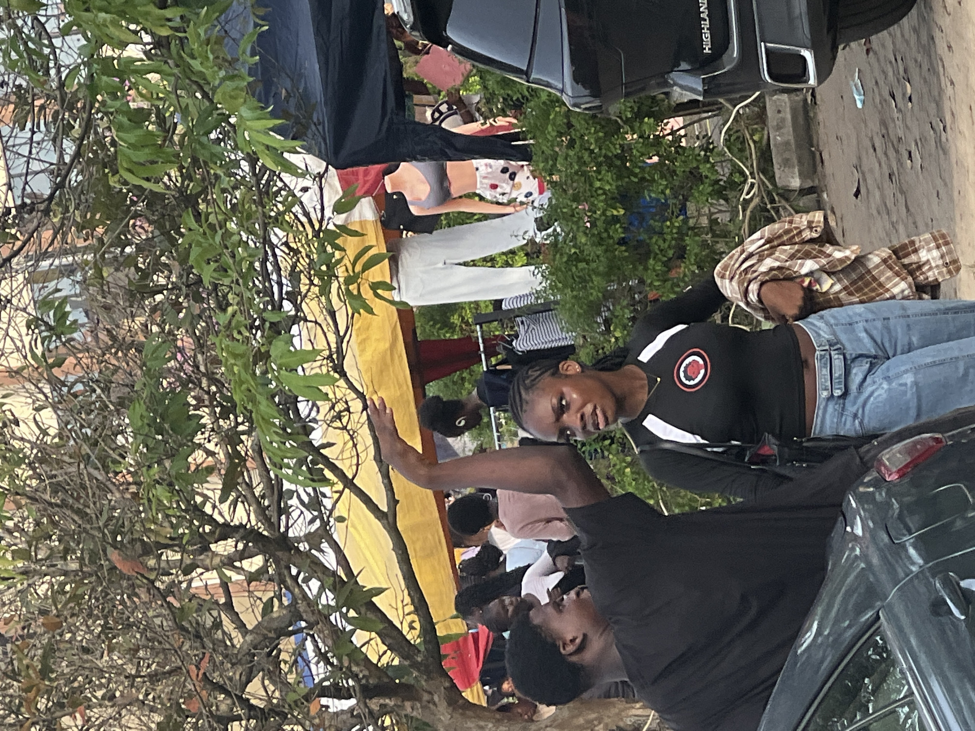 The image shows an outdoor scene likely in Ijanikin, Nigeria. Several people are present, including a young woman in a black shirt with a red logo and light blue jeans, and a young man in a black shirt with his arm raised. They appear to be interacting, possibly talking or gesturing. In the background, more people can be seen gathered near a yellow and red structure, which might be a temporary tent or stall. Clothes are displayed on hangers. A black SUV with "HIGHLAND" visible on its rear is partially in the frame, along with another car. A tree with green leaves is visible on the left side of the image. The environment appears to be a street or roadside setting.