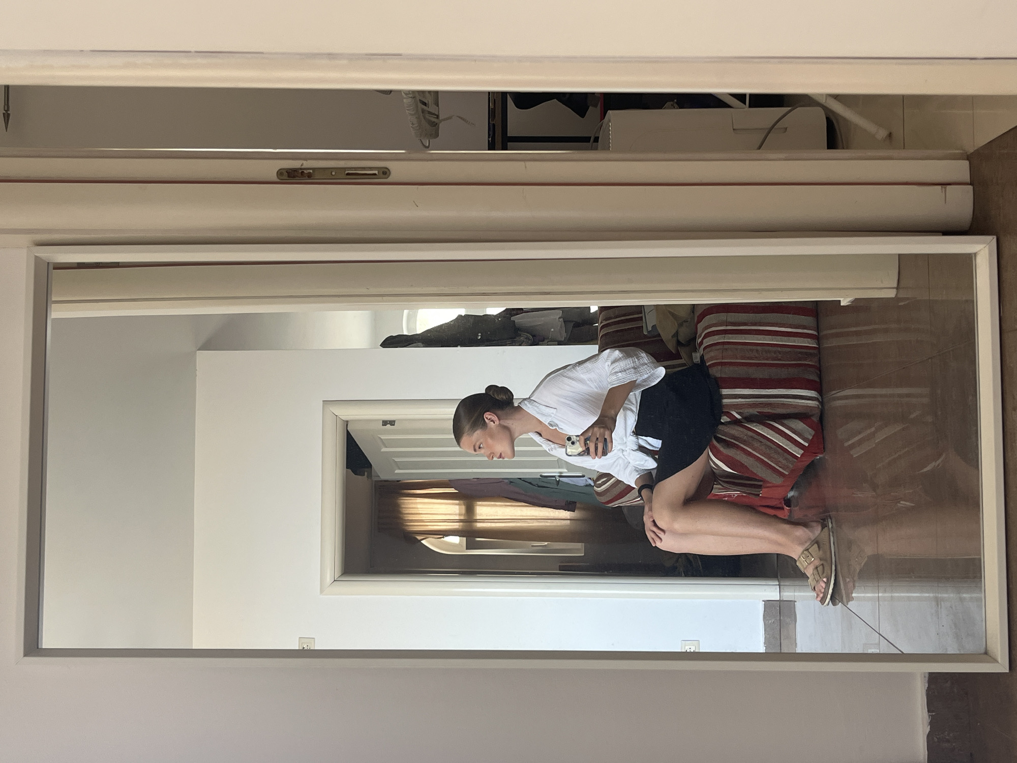 A young woman is seen sitting in a room, reflected in a large mirror. She is wearing a white, short-sleeved button-down shirt tied at the waist and a black skirt. On her feet, she has light brown sandals. Her hair is pulled back into a bun. In her right hand, she holds a smartphone. She is seated on a cushioned stool with a striped pattern in red, beige, and white. The room appears to be a living space with a tiled floor and a doorway in the background leading to another room with curtains and clothing hanging. To the right of the mirror, a doorway frame is visible, with a closed door partially obstructing the view into another area where some furniture and appliances can be seen. The image is taken indoors, likely in a residential setting, potentially an apartment or hotel room, and the style of the doorframe suggests a Mediterranean influence, possibly consistent with locations like Budva, Montenegro, which features a mix of older and newer architecture with similar interior finishes.