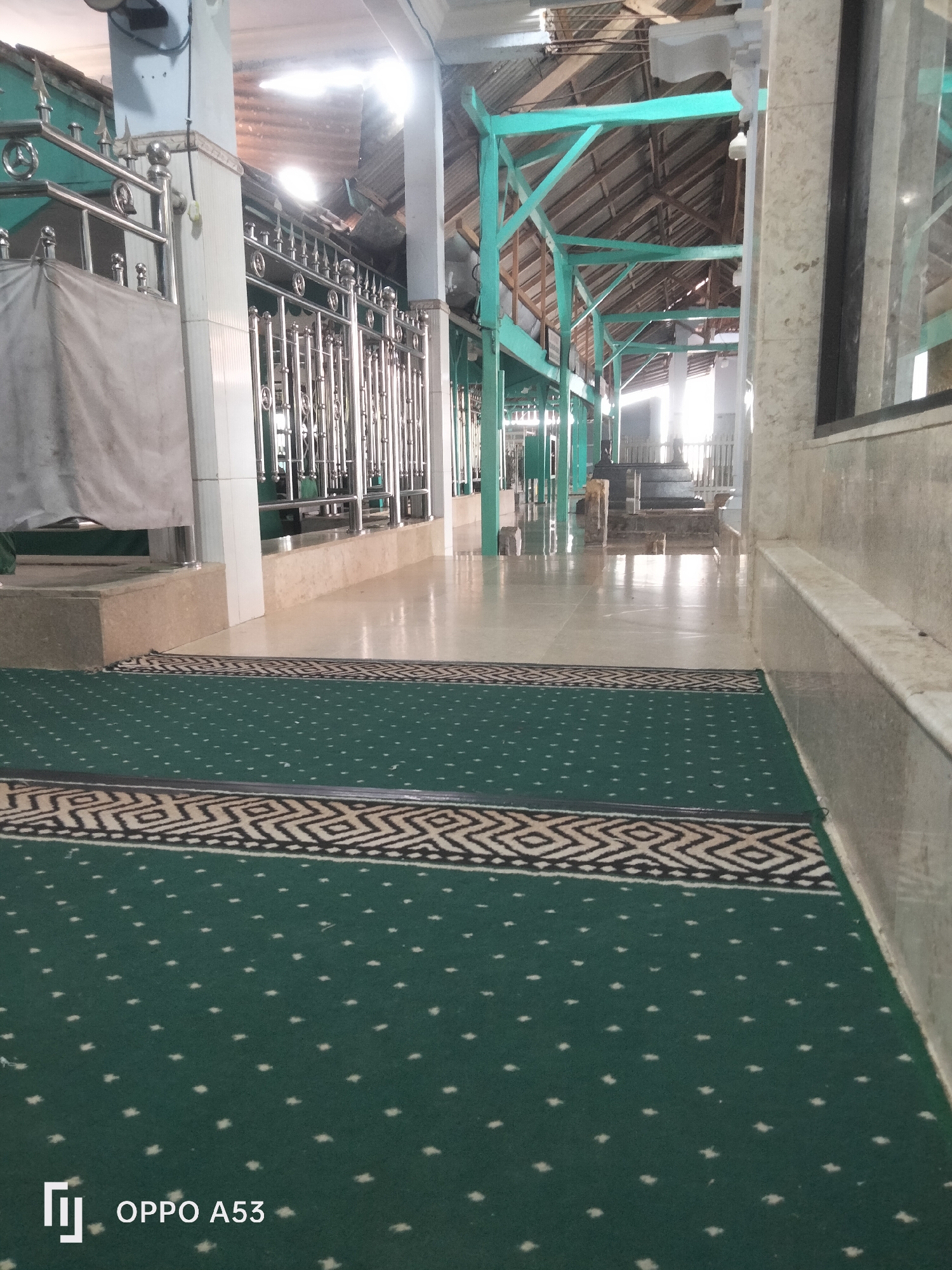 The image shows the interior of a building, likely a mosque, in Badung, Indonesia. In the foreground, there is a green carpet with a repeating pattern of small white diamond shapes and a decorative border with black and white geometric designs. The carpet covers the floor of what appears to be a hall or prayer area. In the midground, the floor is polished and reflects light, suggesting it is made of tile or smooth concrete. A metal railing, likely part of a barrier or enclosure, runs along the left side of the midground, separating it from a lighter-colored tiled wall. Behind the railing, a light-colored cloth or curtain is visible. The background reveals the building's structure, with wooden beams and a corrugated metal roof. The lighting suggests daytime, with sunlight streaming in from the open areas. The overall scene is one of an indoor public space, possibly a place of worship or a memorial site given the presence of a railing and curtain. The "OPPO A53" watermark in the bottom left corner indicates the image was taken with an Oppo A53 smartphone.
