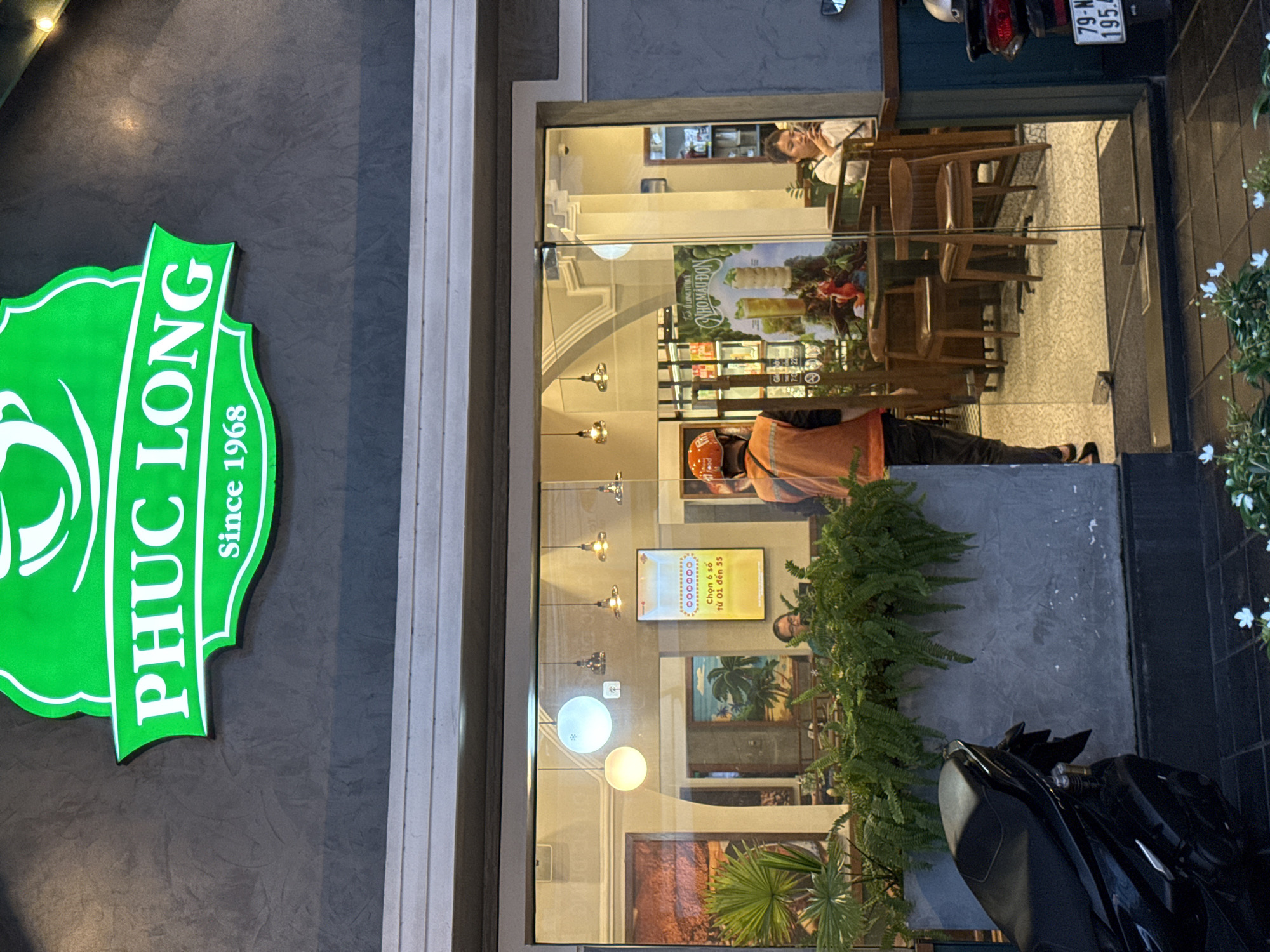 The image shows the exterior of a Phuc Long coffee shop in Nha Trang, Vietnam. The shop's green and white logo is visible at the top of the facade, displaying "PHUC LONG" and "Since 1968." The entrance to the shop features a glass door and large windows, offering a view of the interior. Inside, several tables and chairs are arranged, and plants are visible on the right side of the entrance. A woman is seated at a table on the right, looking at her phone. A person wearing an orange vest and helmet, likely an employee or delivery person, stands near the entrance. A motorcycle is parked to the left of the entrance, and another motorcycle's license plate, reading "79-N 195.4", is visible on the right side of the image. The facade is gray and textured, with white trim around the windows and entrance. The time of day appears to be evening or late afternoon, as indicated by the lighting.