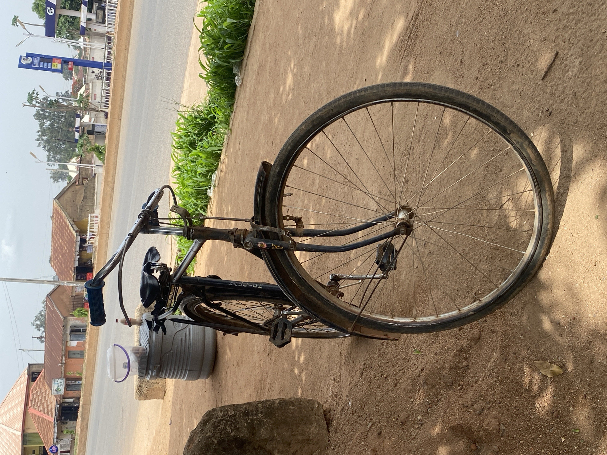 A black bicycle stands on a dirt shoulder beside a paved road in Bukuru, Nigeria. The front wheel is prominent, with thin, rusted spokes. A large gray cooler sits behind the seat of the bike, with a plastic bucket placed on top. In the background, buildings with rusty, orange roofs and a gas station can be seen across the road. Green vegetation lines the edge of the shoulder. The image is oriented sideways, requiring rotation for proper viewing.
