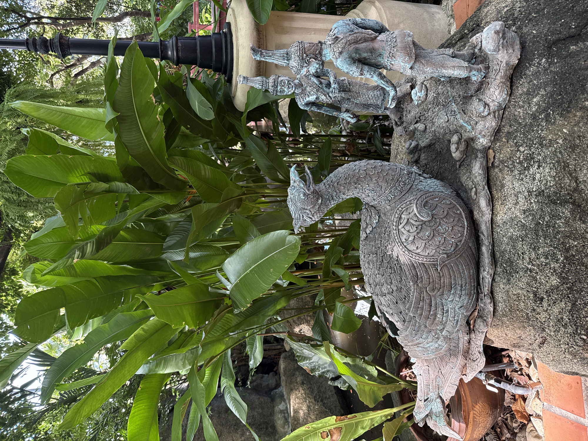 The image depicts a garden setting in Samrong Nuea Subdistrict Municipality, Thailand. A bronze-colored statue of a bird, resembling a peacock, is positioned in the foreground. The bird is highly detailed, with intricate patterns on its feathers. Behind the peacock statue, there are two bronze-colored statues of male figures wearing traditional Thai headdresses. These figures appear to be positioned on a rock or stone structure. Lush green foliage, including large, broad leaves, fills the background, obscuring some of the statues and structures. A black lamppost is visible among the leaves. The statues are situated on a textured stone surface with a mix of natural elements like rocks and plants. The overall impression is that of a serene, landscaped garden with culturally significant sculptures.