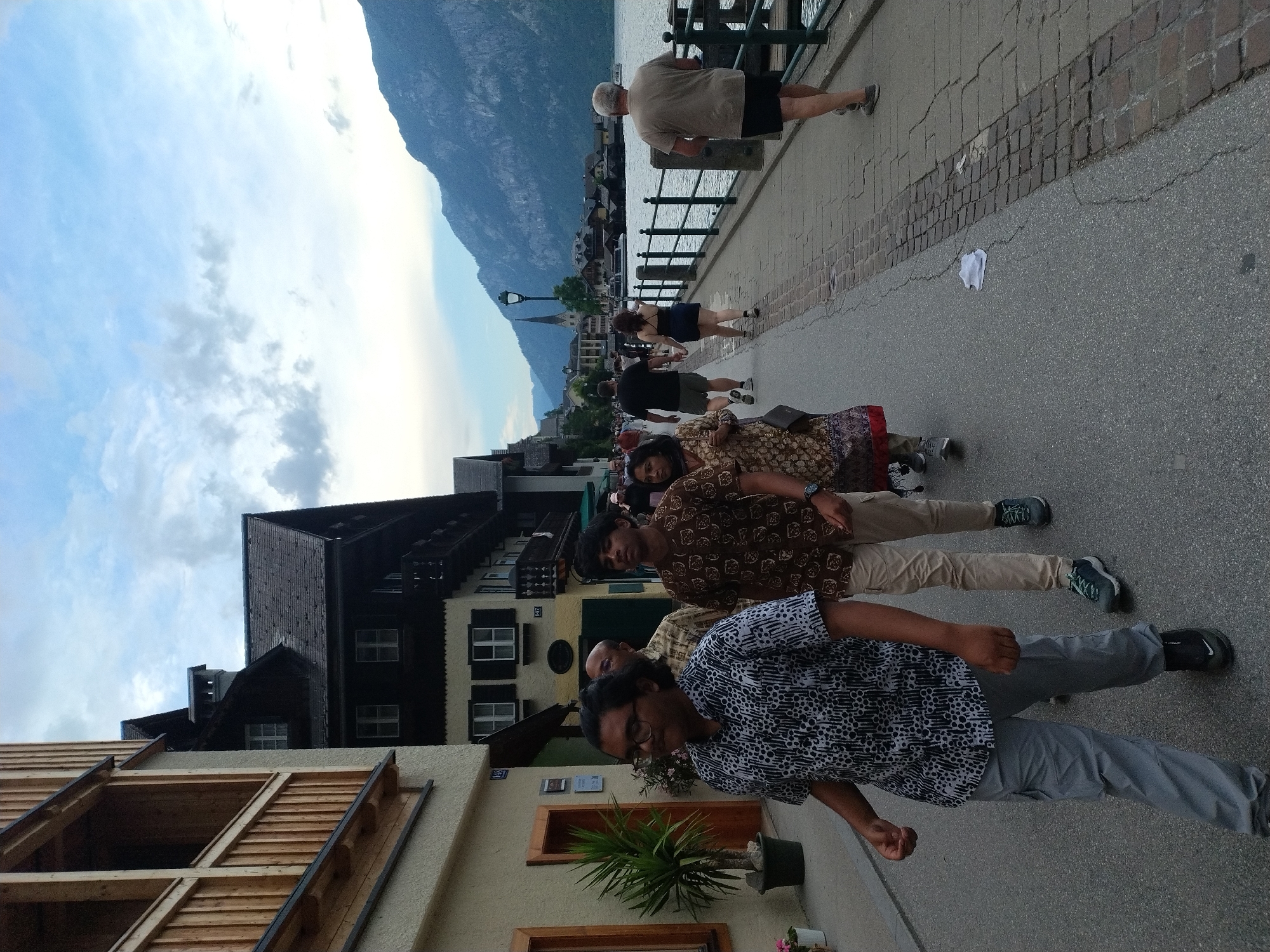 A group of people are walking along a cobblestone pathway next to a lake in Hallstatt, Austria. The pathway is lined with a black metal railing. In the background, a body of water is visible, with a mountain range in the distance. The buildings on the left are traditional Austrian architecture. A young man in a patterned shirt and grey pants is in the foreground, walking towards the viewer. He has dark, curly hair and is wearing glasses. Behind him, a man in a patterned shirt and khaki pants is also walking. Further back, a woman in a colorful patterned skirt is visible. On the right side of the pathway, a man in a light brown t-shirt and black shorts is walking away from the camera. The sky is cloudy with patches of blue.