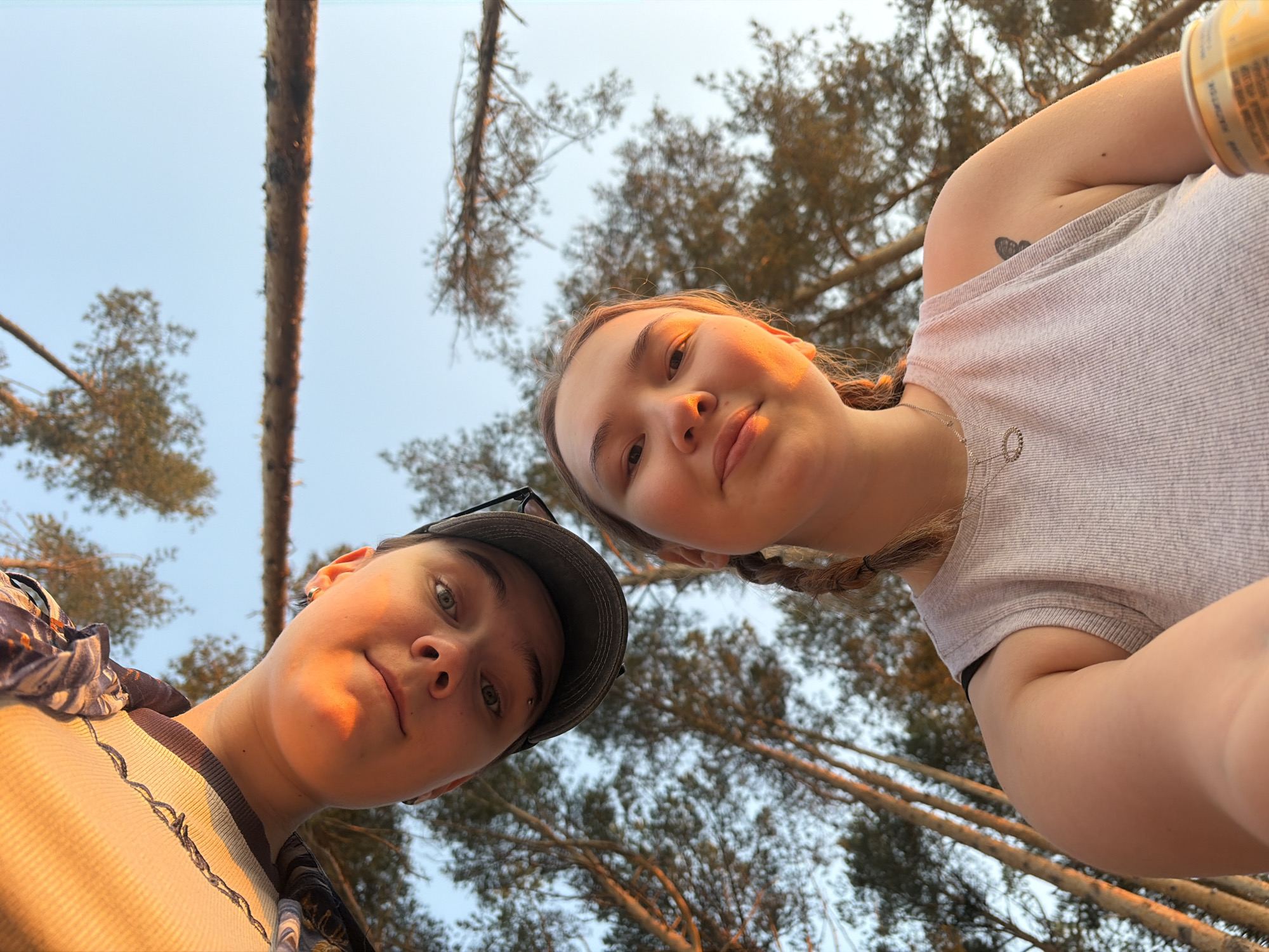 Two individuals, a fair-skinned person wearing a baseball cap and a fair-skinned person with braided hair, are positioned in the foreground, looking down towards the camera. Behind them, a wooded area is visible, with tall trees reaching upwards towards a pale sky. The angle suggests a ground-level perspective looking up. A can is visible in the upper right corner. The location is Chaikovsky, Russia, based on the available contextual information.
