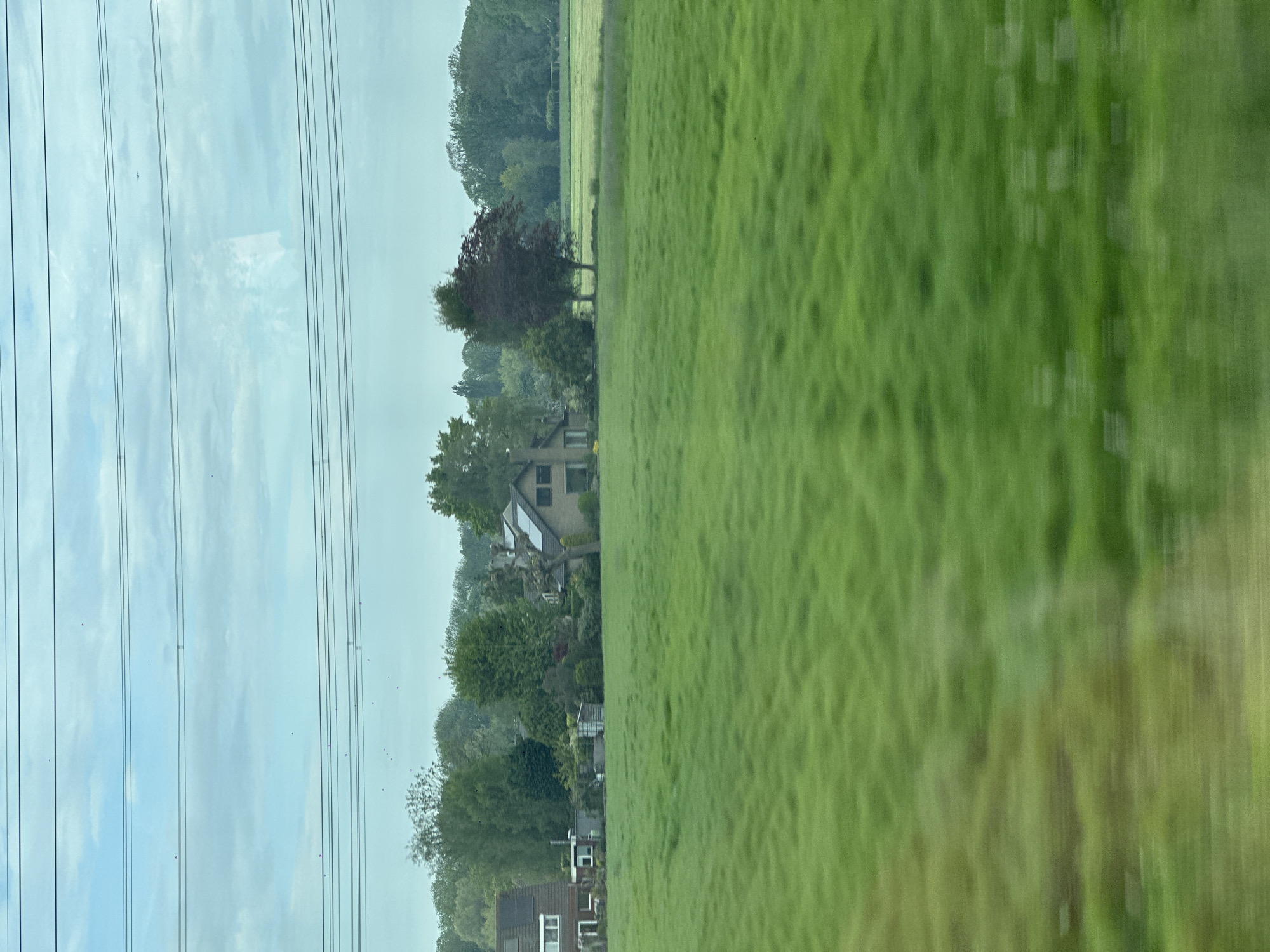 The image presents a view from a moving train, likely in a rural setting. A vast green field dominates the right side of the frame, its texture blurred due to the motion. On the left side, power lines extend across the sky, suggesting an industrialized area. Beyond the power lines, there are a few houses nestled among trees, partially obscured. There is no evidence that this is Loenersloot, Netherlands. No individuals are visible, the focus being on the landscape and infrastructure. The overall impression is one of a journey through a mixed environment - rural and developed.