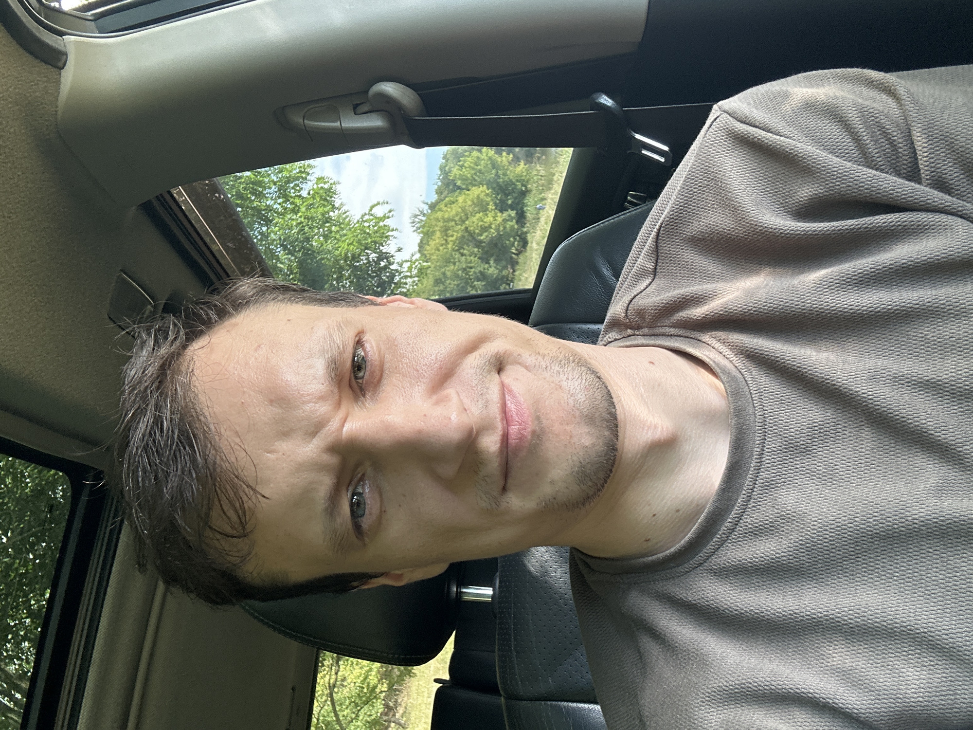 A man is seen from the chest up, inside a vehicle. He is looking at the camera with a slight frown and his eyebrows are raised. The man has short, dark hair and light-colored eyes. He is wearing a tan t-shirt. The interior of the car is visible behind him, including a black leather seat and part of the dashboard. A sunroof is open above, showing green foliage. The background outside the car window is also filled with trees and a cloudy sky. The scene appears to be outdoors in a natural setting, possibly a rural area. There is no indication of Makiv, Ukraine.