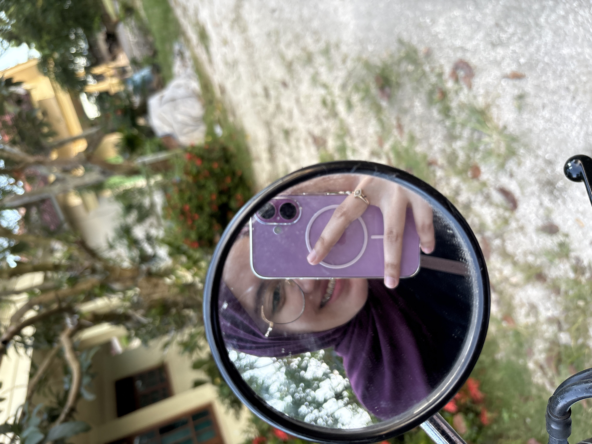A person is taking a selfie in a circular mirror attached to what appears to be a vehicle, likely a motorcycle, in an outdoor setting. The person reflected in the mirror is wearing glasses and a purple hijab, holding a purple cell phone to take the photo. The background visible through the mirror includes a blurred view of greenery, a building, and a person in white clothing tending to plants. The ground appears to be covered in loose gravel or dirt. The location is likely Gadingrejo, Indonesia, based on the prompt.