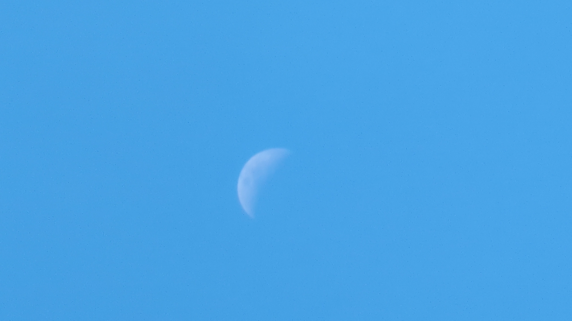 The image shows a partial view of the moon against a clear blue sky. The moon is illuminated on the right side, with a crescent shape visible. The sky is uniformly blue with a slight texture of small, speckled dots that could be noise from the image capture or atmospheric particles. There are no discernable landmarks or elements that would suggest a specific location in Kozare, Bulgaria, or any other city. The scene is entirely natural, focusing on the celestial body and the sky.