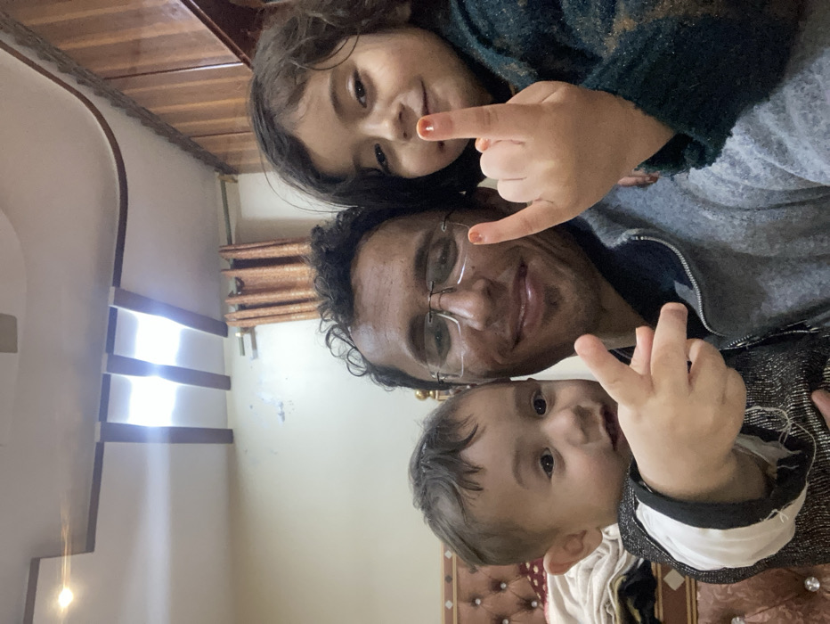 A selfie-style photo shows a man, likely a father, with two young children. The man wears glasses and smiles. A girl is at the top of the frame, flashing a "rock on" hand gesture. A young boy at the bottom of the frame makes a peace sign with his fingers. The background shows a room interior with decorative elements, including curtains and a cushioned headboard. The photo is taken at an angle, with the subjects' faces close together.