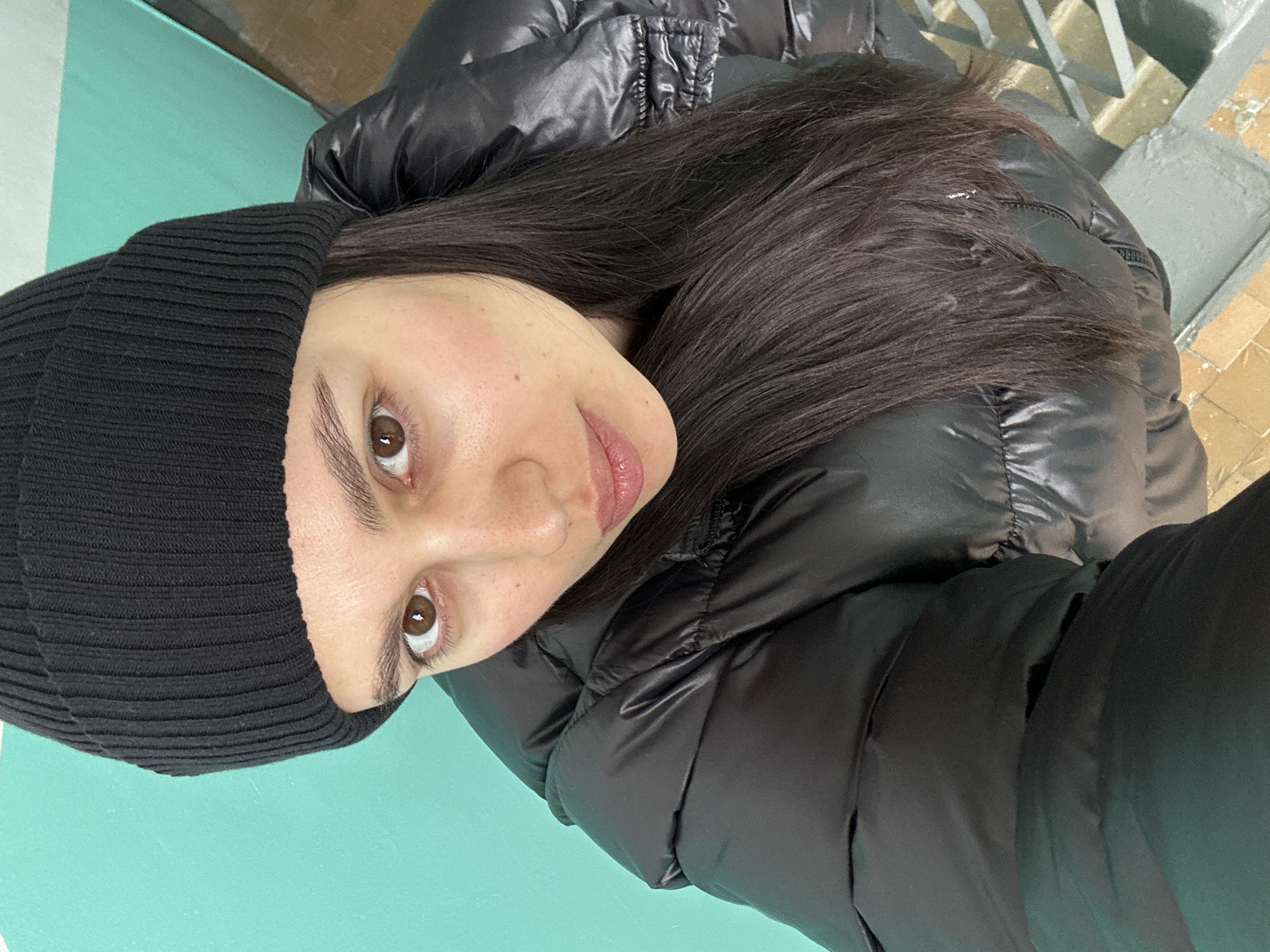 A young woman is depicted in a selfie, seemingly taken outdoors. She wears a black beanie and a black, puffy jacket. Her hair is dark and appears to be straight. The background is blurry but suggests an urban environment, possibly in Chaikovsky, Russia. Visible elements include a green/blue painted surface, likely a wall or floor, and a glimpse of stairs or a structure, suggesting an outdoor stairwell.