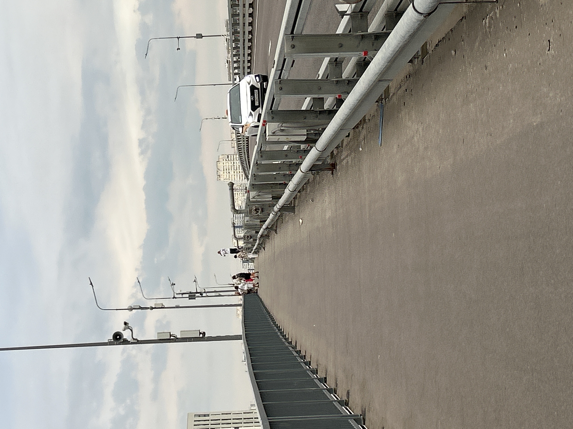 The image shows a pedestrian walkway on a bridge in an urban environment. The walkway is made of concrete and is bordered by a metal railing on the left and the road on the right. Several streetlights with cameras and speakers are visible along the walkway. In the distance, a white taxi is driving on the road. A group of people are standing on the walkway, and one person is standing on the railing in the distance. Buildings are visible in the background. The sky is overcast.