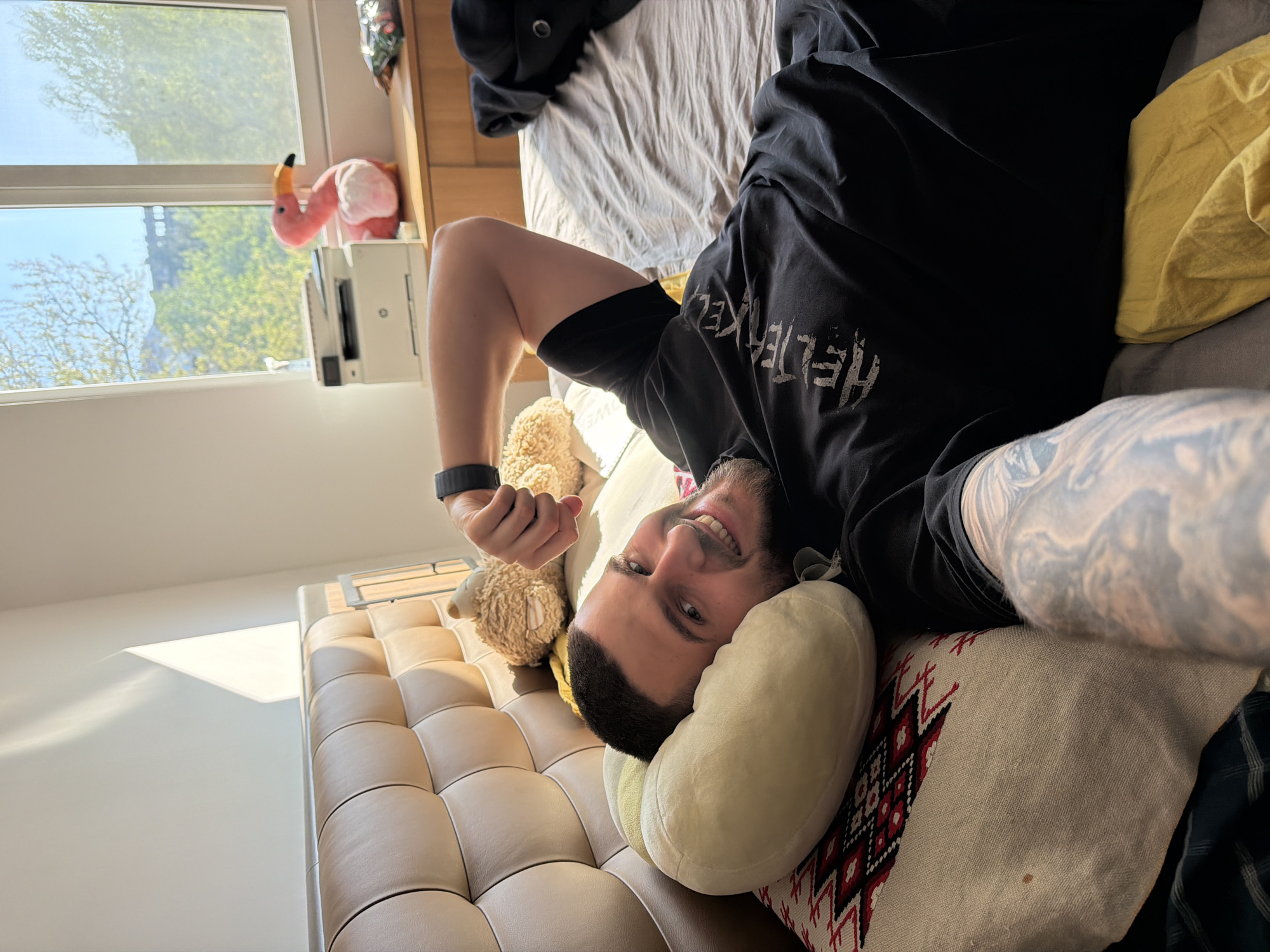 A man is lying on his back in what appears to be a bedroom or living area. He is fair-skinned with dark hair and a beard, wearing a black t-shirt, a wristband, and has visible tattoos on his arm. He is smiling. His head rests on a light-colored neck pillow, with a decorative cushion partially visible underneath. To his left, there is a beige tufted sofa or bench with two small teddy bear stuffed animals.
In the background, a window reveals an outdoor view with buildings and trees, indicating a daytime scene. A pink flamingo toy and what looks like a printer are on a surface near the window. A bed with a grey blanket is also visible in the background. The image does not provide information that confirms the location as the city of Sofia, Bulgaria.
