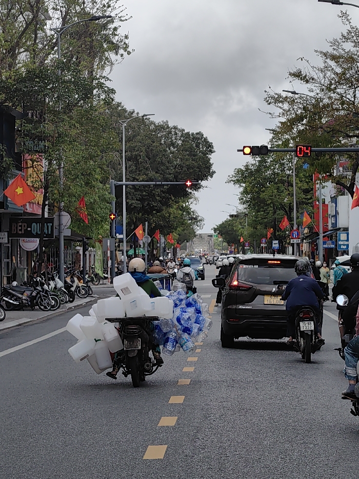 The image depicts a busy street scene in Thuận Hóa Ward, Vietnam, under an overcast sky. The asphalt road features dashed yellow lane markings. In the foreground, a person wearing a light-colored helmet and a dark jacket is operating a dark-colored motorcycle, positioned in the left lane and facing away from the viewer. This motorcycle is heavily loaded with numerous white, opaque plastic containers secured to the left side of its rear and multiple bundles of clear plastic water bottles secured to the right side.
Ahead of this motorcycle, in the right lane, a black Mitsubishi Expander SUV (license plate 23H 161.00) is visible, followed closely by another person on a motorcycle, wearing a dark helmet and a blue shirt (license plate 25-NB T332). Overhead, a traffic signal to the right displays a yellow light and a red digital countdown timer showing "04". Further down the street, another traffic light indicates red.
Buildings line both sides of the street, with shops visible on the left, displaying signage including "BẾP-QUẠT" and "MÓN ĂN." Numerous small Vietnamese flags (red with a yellow star) are displayed along the street, attached to poles and buildings. Green trees with dense foliage line the sidewalks and extend over the roadway. Multiple streetlights with modern LED fixtures are present. A row of parked motorcycles is visible along the left sidewalk, and additional vehicles and pedestrians are discernible in the background.