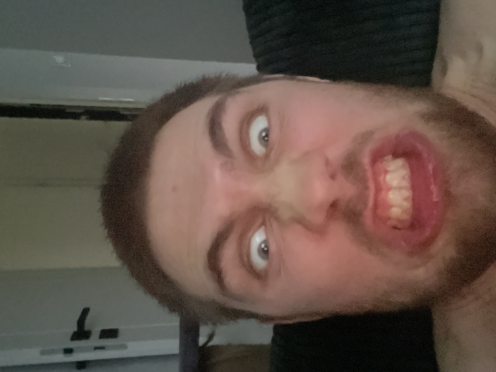 A close-up, low-angle selfie of a man with light brown hair and a short beard is captured. He is making a comical facial expression with his eyes wide open and his teeth bared in a grimace. His mouth is open, revealing his teeth. The background is dimly lit, with a door handle visible on the left side of the frame. There are no discernible landmarks or features that would indicate a specific location within Pruszcz Gdański, Poland.