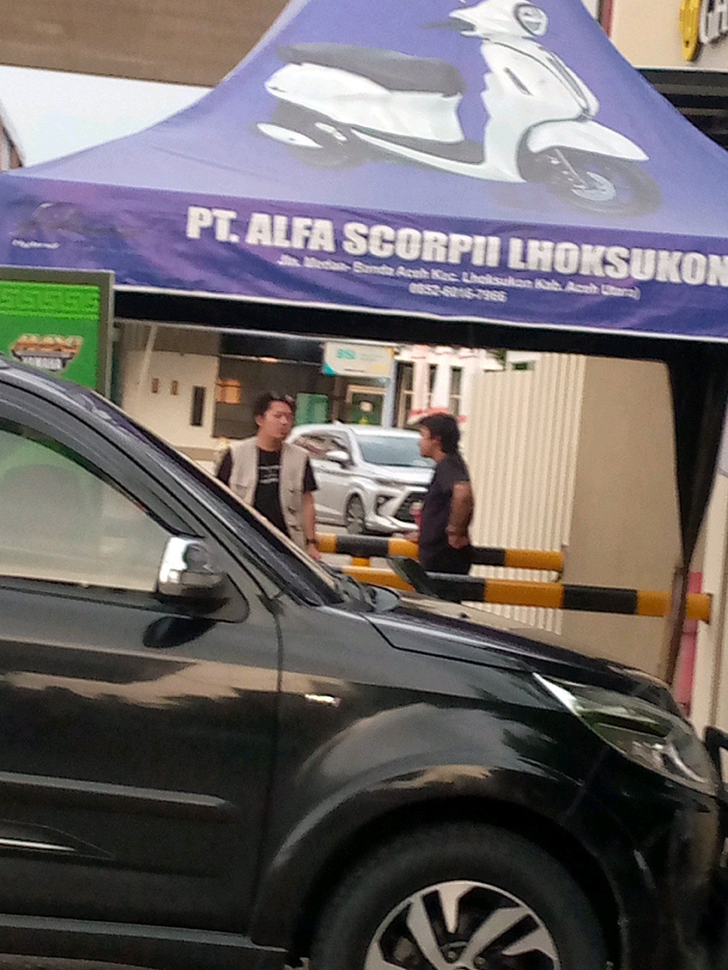 The image depicts a scene in Keudeteupinpunti, Indonesia, likely outside a business. A dark-colored car is in the foreground, partially obscuring a tent with a purple banner. The banner displays white text that reads "PT. ALFA SCORPIII LHOKSUKON" and smaller text below it indicating an address and phone number. A white scooter is illustrated on the purple banner. Two men are visible in the background, standing near the tent. One man, wearing a tan vest over a dark shirt, appears to be looking to his right. The other man, wearing a dark shirt, is seen from the side, facing away from the camera. Behind the men, a white car is parked, and a striped barrier is visible. The environment suggests a commercial area, possibly a dealership or a promotional event.