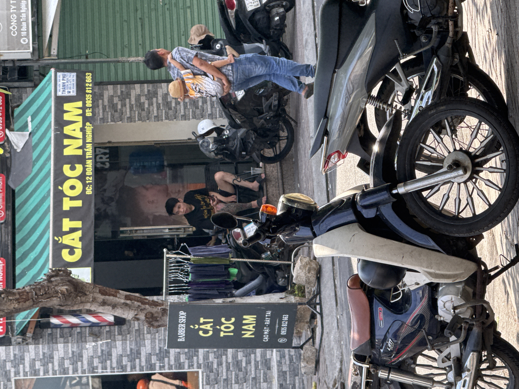 The image depicts a street scene in Bac Nha Trang Ward, Vietnam, focusing on a barbershop. A black sign with "BARBER SHOP" in white text and "CẮT TÓC NAM" (Men's Haircut) in yellow text is prominent in the foreground. Below this, smaller text reads "CAO MẶT - RÂY TAI" (Shave Face - Ear Wax) and a phone number, "0935 812 663". To the right of the sign, a rack of men's clothing hangs outdoors.
A young man is seated on a step outside the barbershop, looking at his phone. Behind him, a man carries a child on his back, walking away from the camera. Several motorcycles are parked on the street. The barbershop's facade features a green and yellow striped awning with the text "CẮT TÓC NAM" in large yellow letters. A smaller sign to the right of the awning provides a phone number and address details. The street appears to be paved, with some debris and rocks on the ground. The overall environment suggests an urban setting.